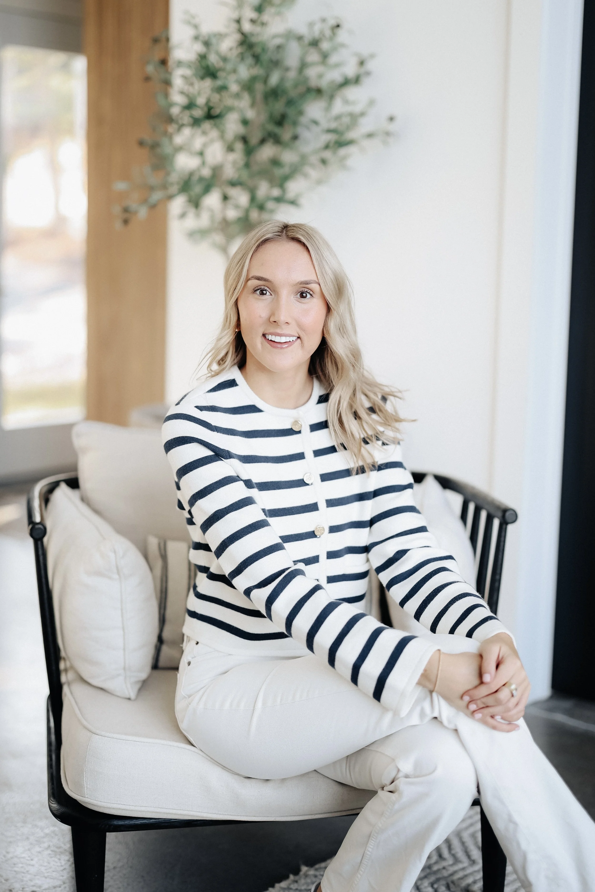 A woman with long blonde hair, wearing a white and navy striped sweater and beige pants, sitting on a white cushioned chair with pillows, smiling in a bright, modern room with a plant in the background.