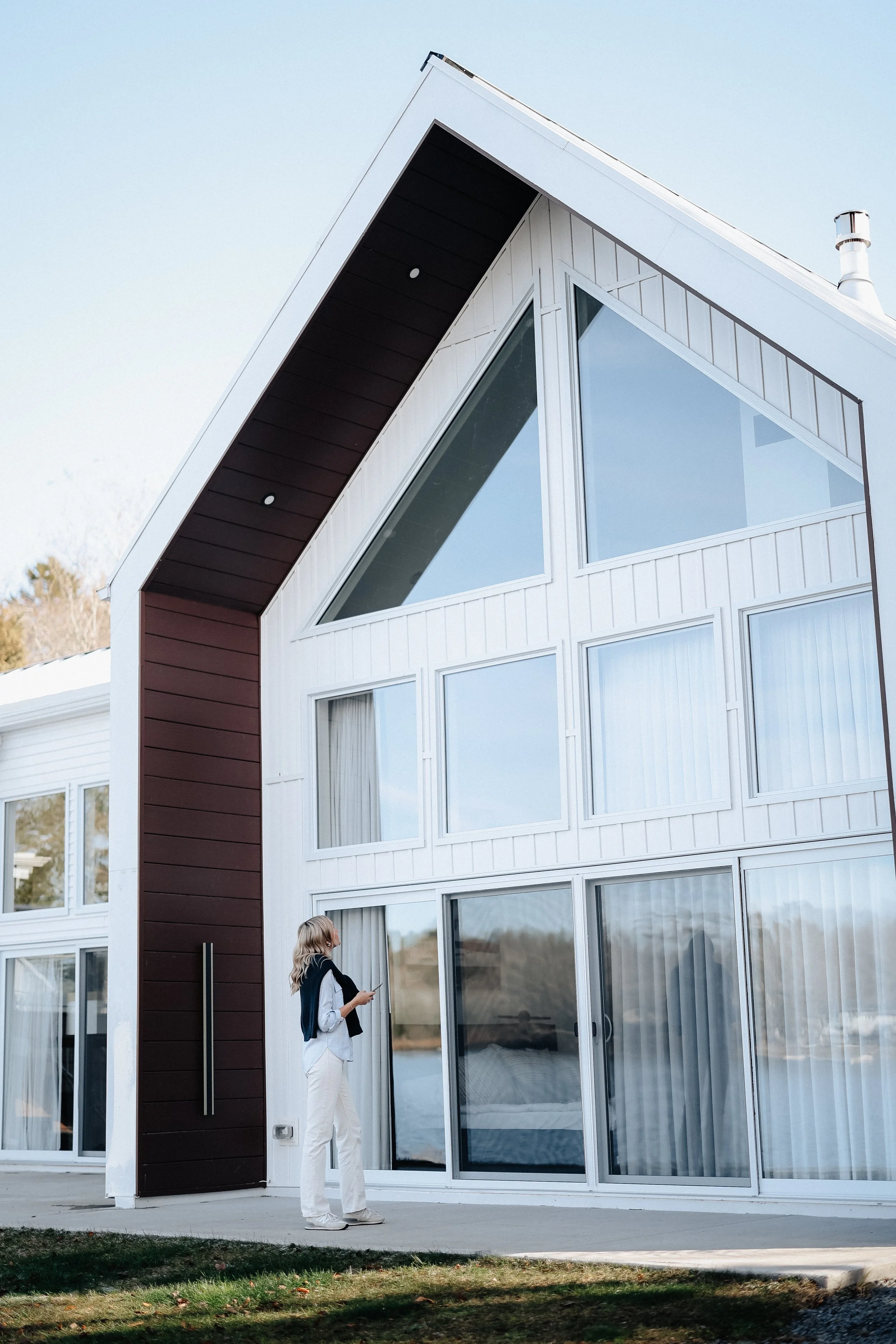 A woman standing in front of a modern house with large windows and a sloped roof, holding a phone and looking at the house.