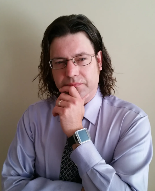 A man with long, dark, wavy hair and glasses, wearing a light purple dress shirt and a patterned tie, standing against a plain beige wall, with his hand on his chin, wearing a smartwatch.