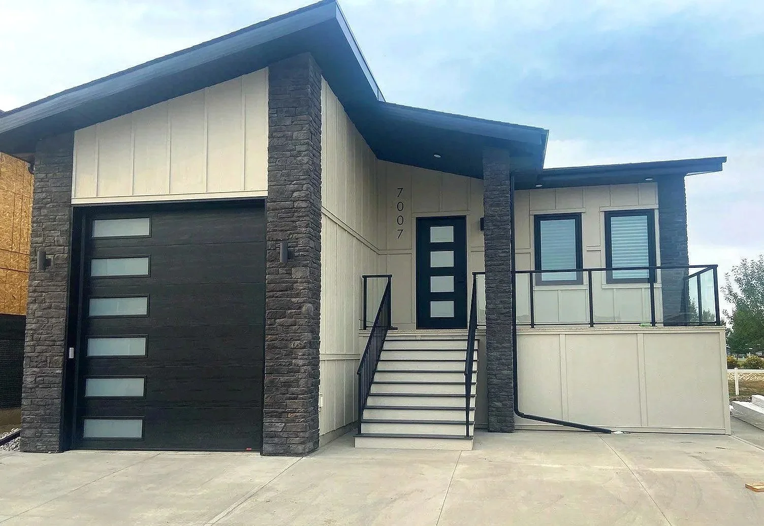 Wicked Maple Design - Modern two-story house with a black garage door, a staircase leading to the front door, and a balcony with a glass railing. The house features black framing around the windows and door, with gray stone accents on the pillars.