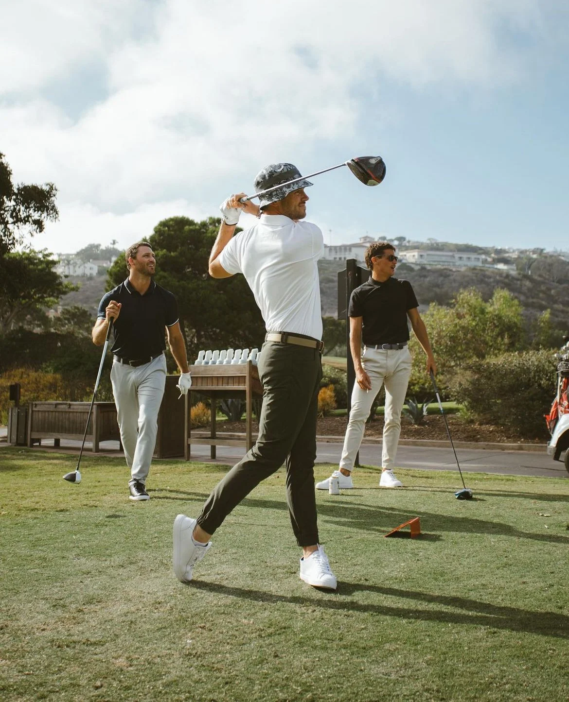 Three men playing golf on a golf course, with one swinging a club and two others watching, surrounded by trees and hills in the background.