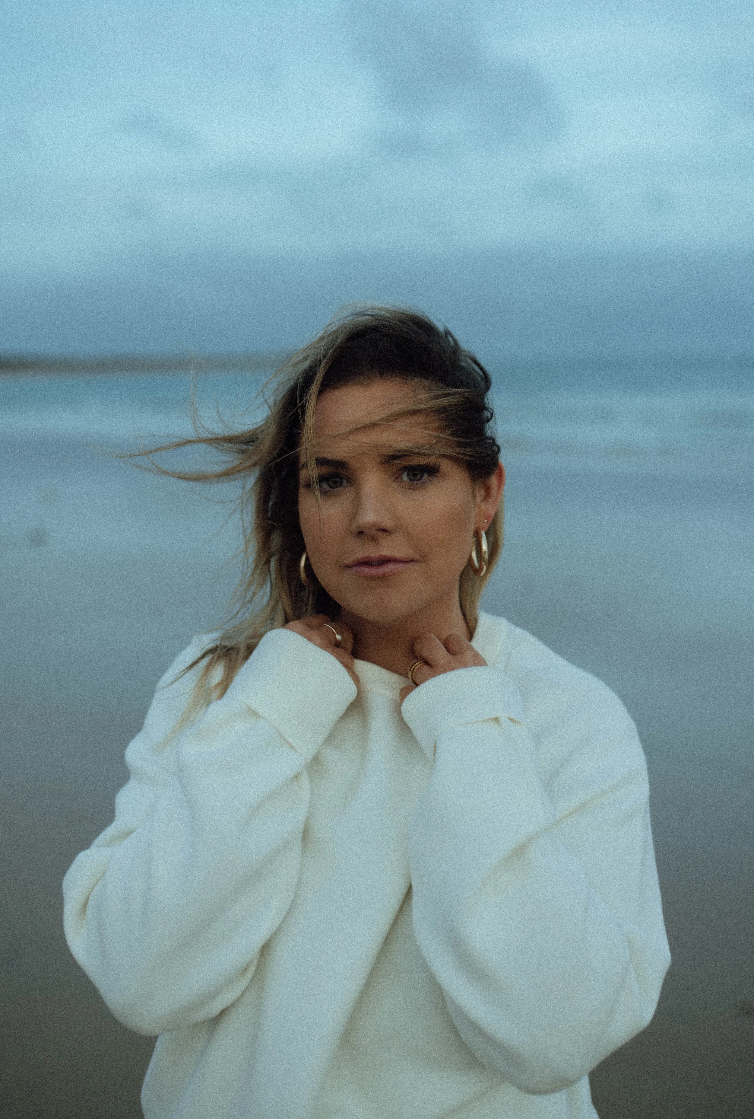 A young woman with blonde hair and hoop earrings, wearing a white sweatshirt, standing outdoors near the water with a cloudy sky in the background.