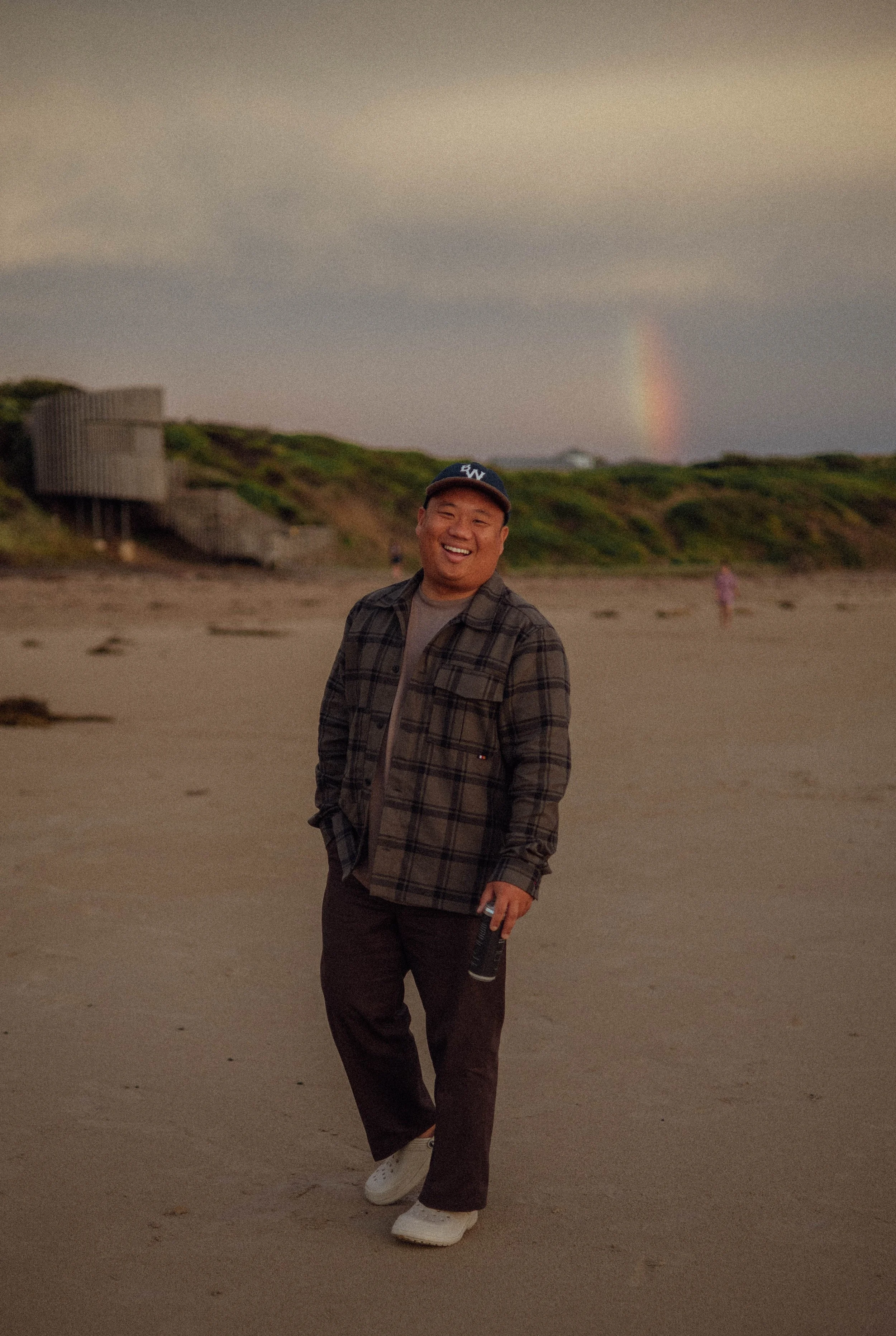 A person smiling and walking on a beach during sunset, with a rainbow in the background and a wooden structure on a hill.
