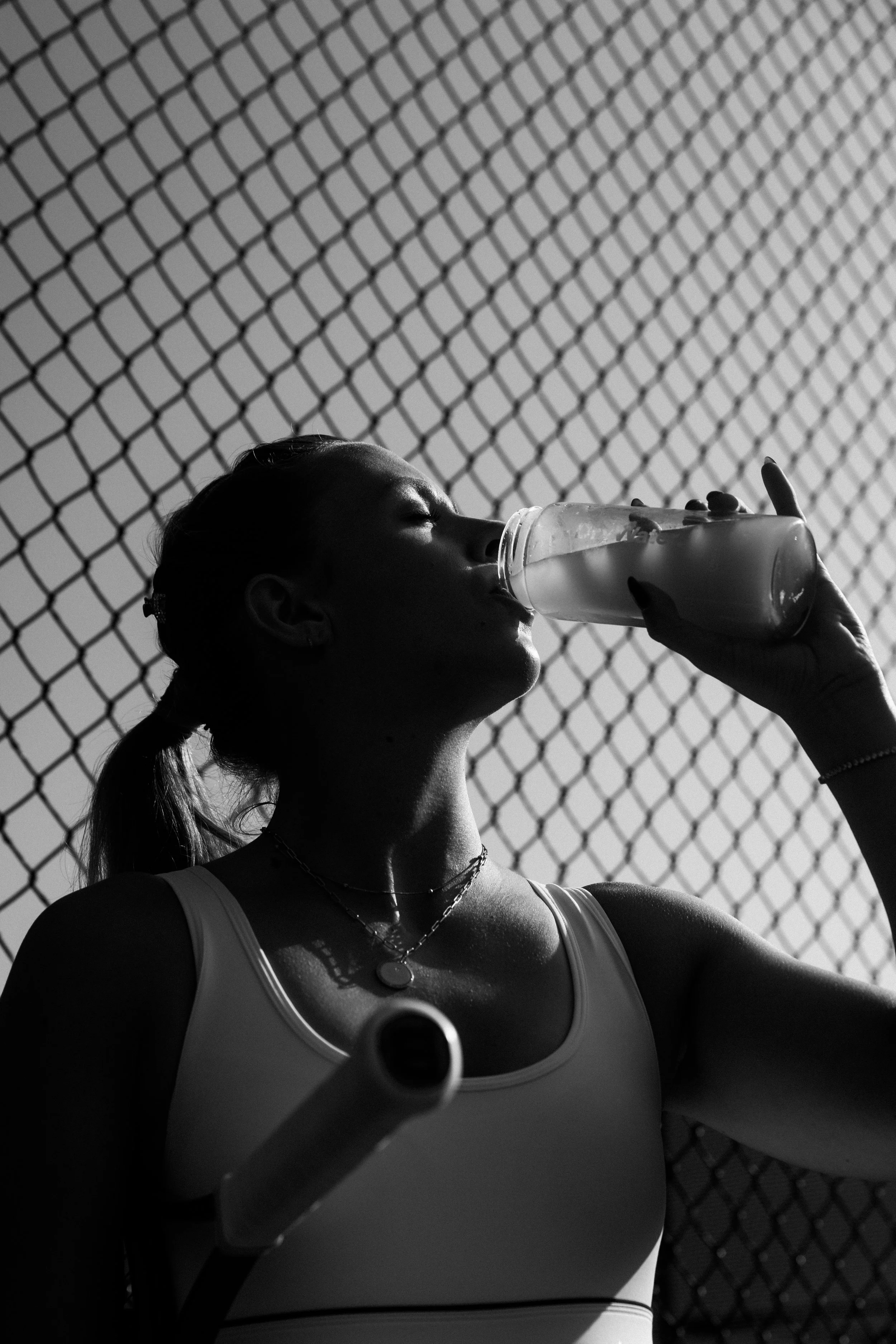 A woman with braided hair drinking water from a plastic bottle, wearing a sports top, necklaces, and earrings, in front of a chain-link fence in sunlight.