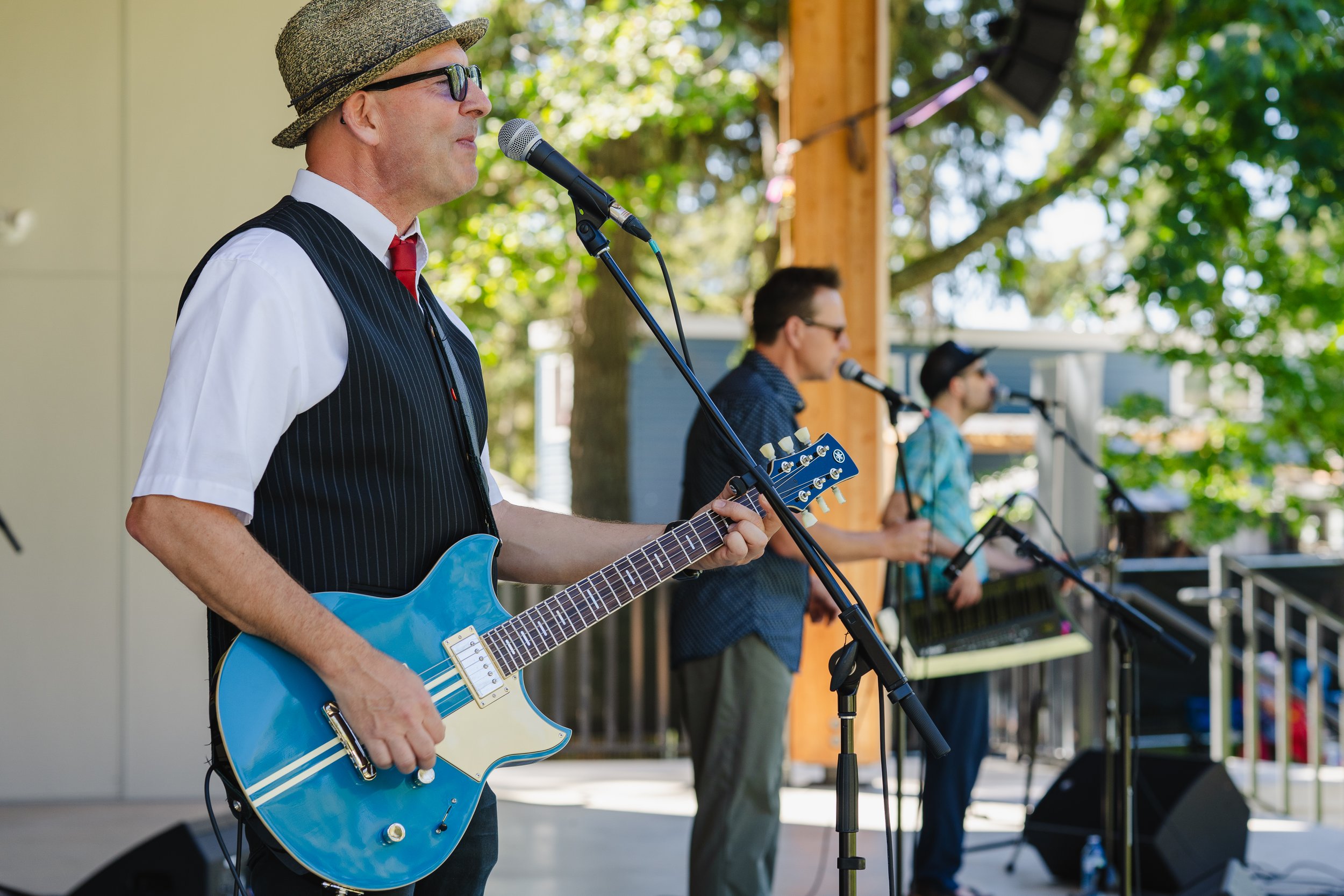 Three musicians perform on an outdoor stage, with the foreground artist playing a blue electric guitar and wearing a hat and sunglasses; two others sing into microphones behind him.