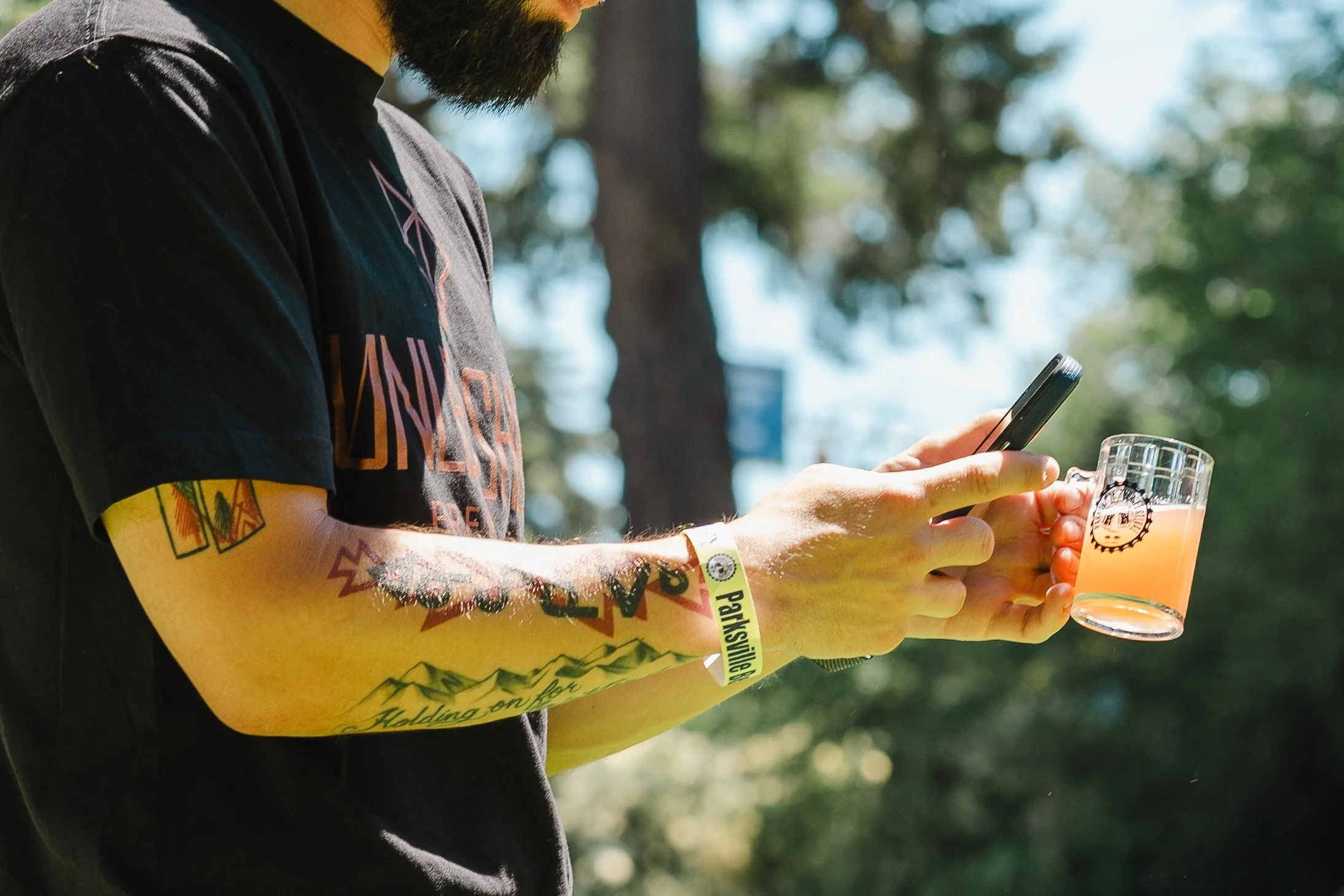 Person holding a small mug of orange drink and a smartphone, wearing a bracelet and a black T-shirt, with tattoos visible on arms, outdoors.
