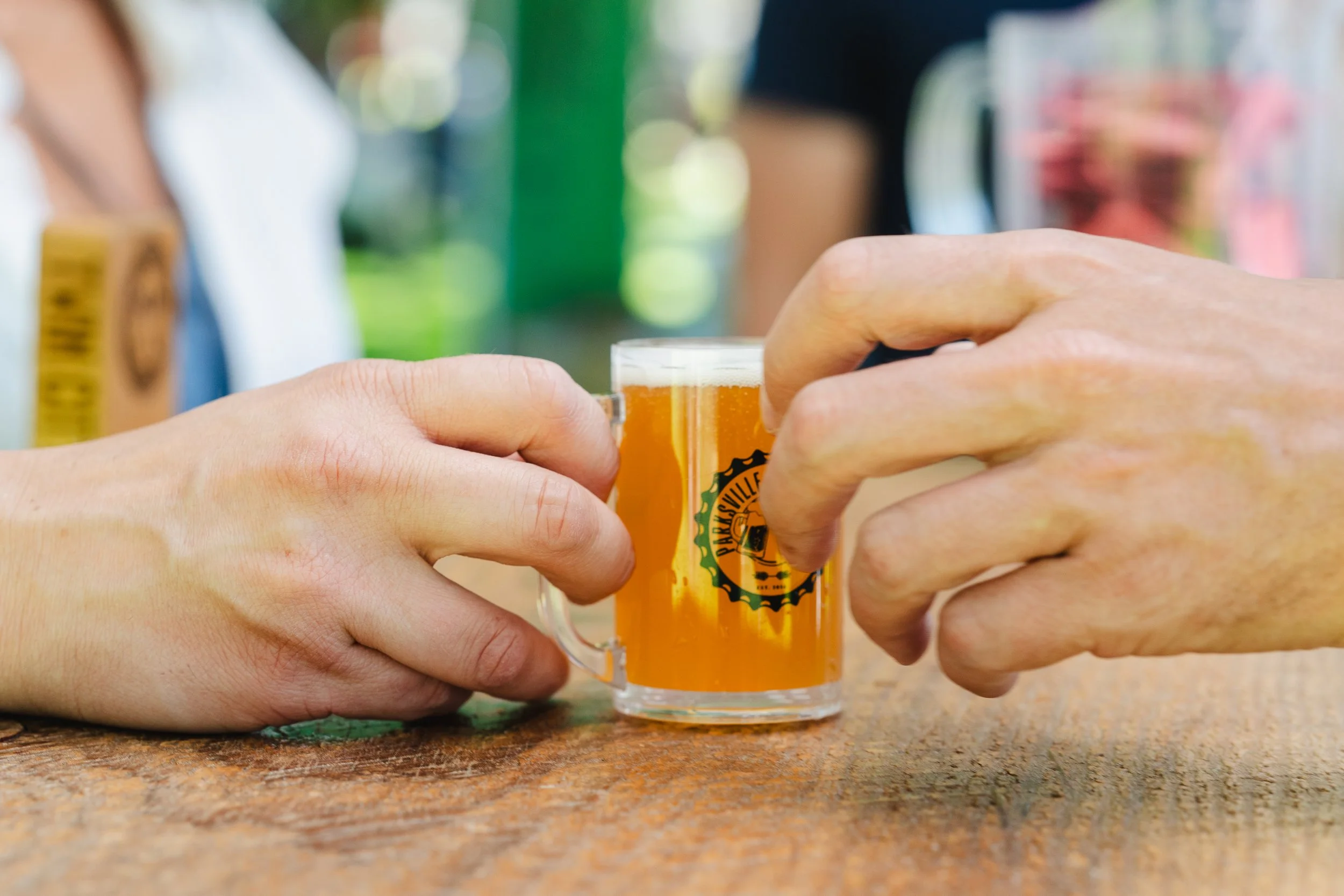 Two people holding a small glass of beer on a wooden table.