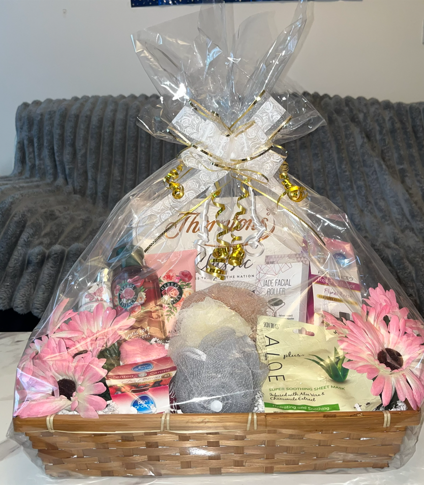 A gift basket wrapped in clear plastic, containing skincare products, a bath sponge, and pink flowers, decorated with gold curly ribbon and a gift tag.