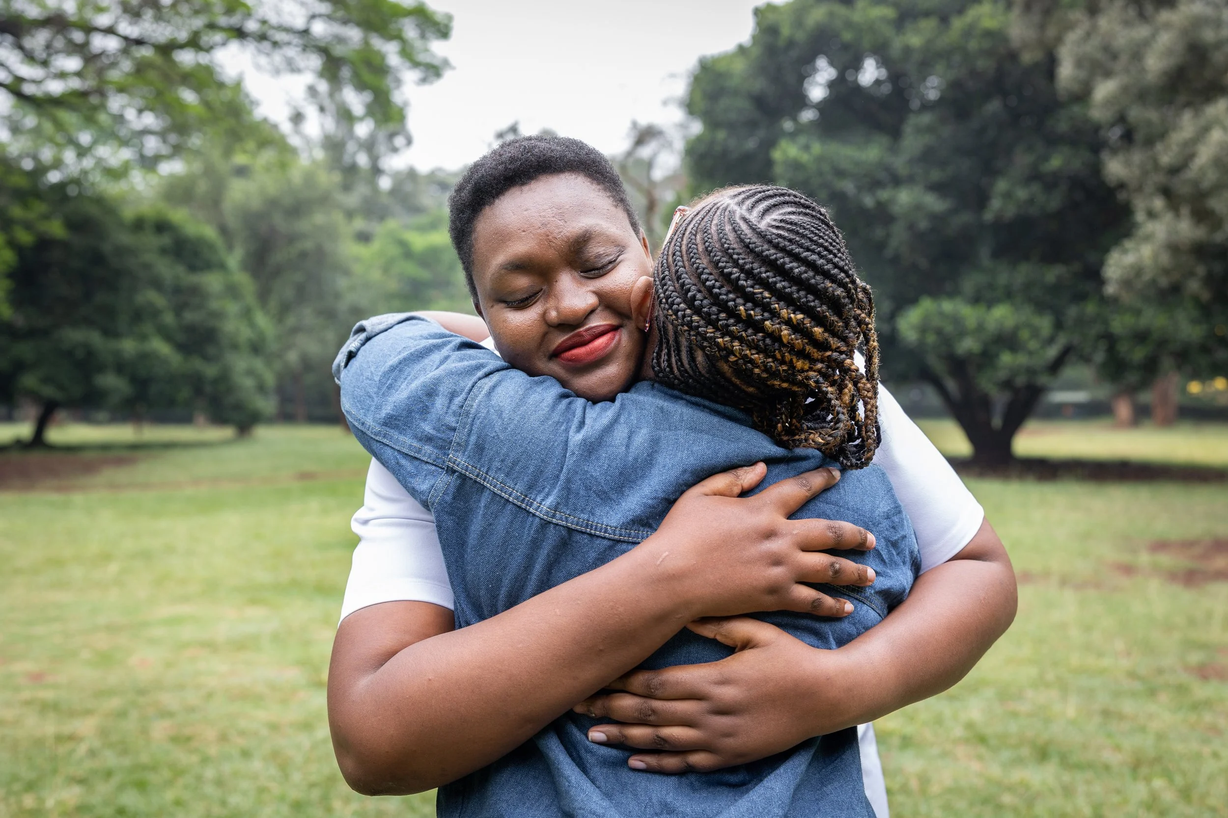 Two people hugging outdoors in a park, with trees and grass in the background. One woman is facing the camera with her eyes closed and a smile, while the other has her back to the camera.