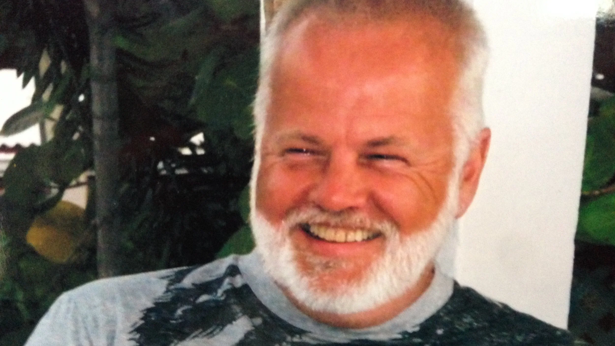 Close-up of a smiling man with gray hair and a beard, wearing a black and white patterned shirt, in front of green plant leaves.
