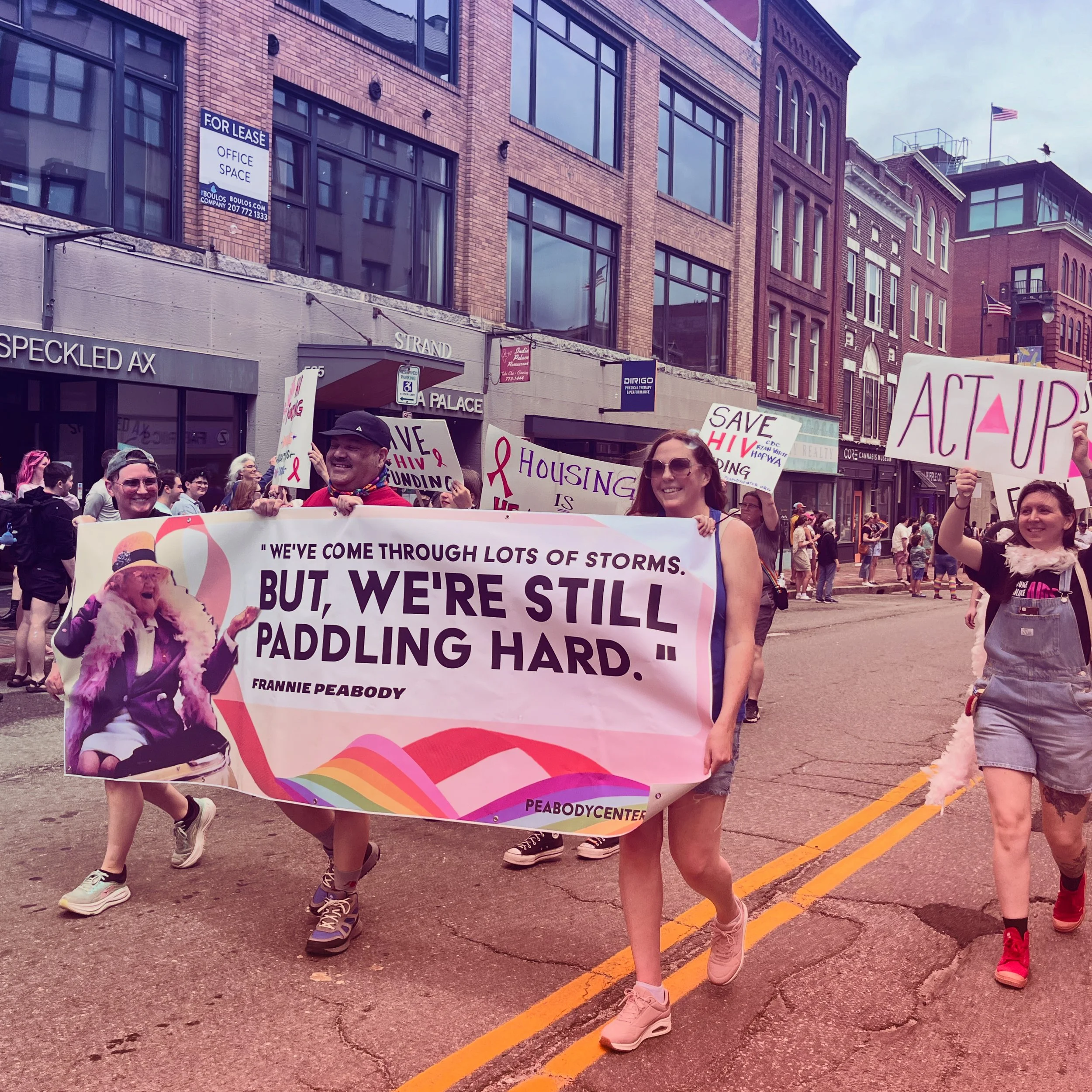 A diverse group of people participating in a pride parade marching down city streets, holding signs supporting HIV awareness and housing rights, with buildings in the background.