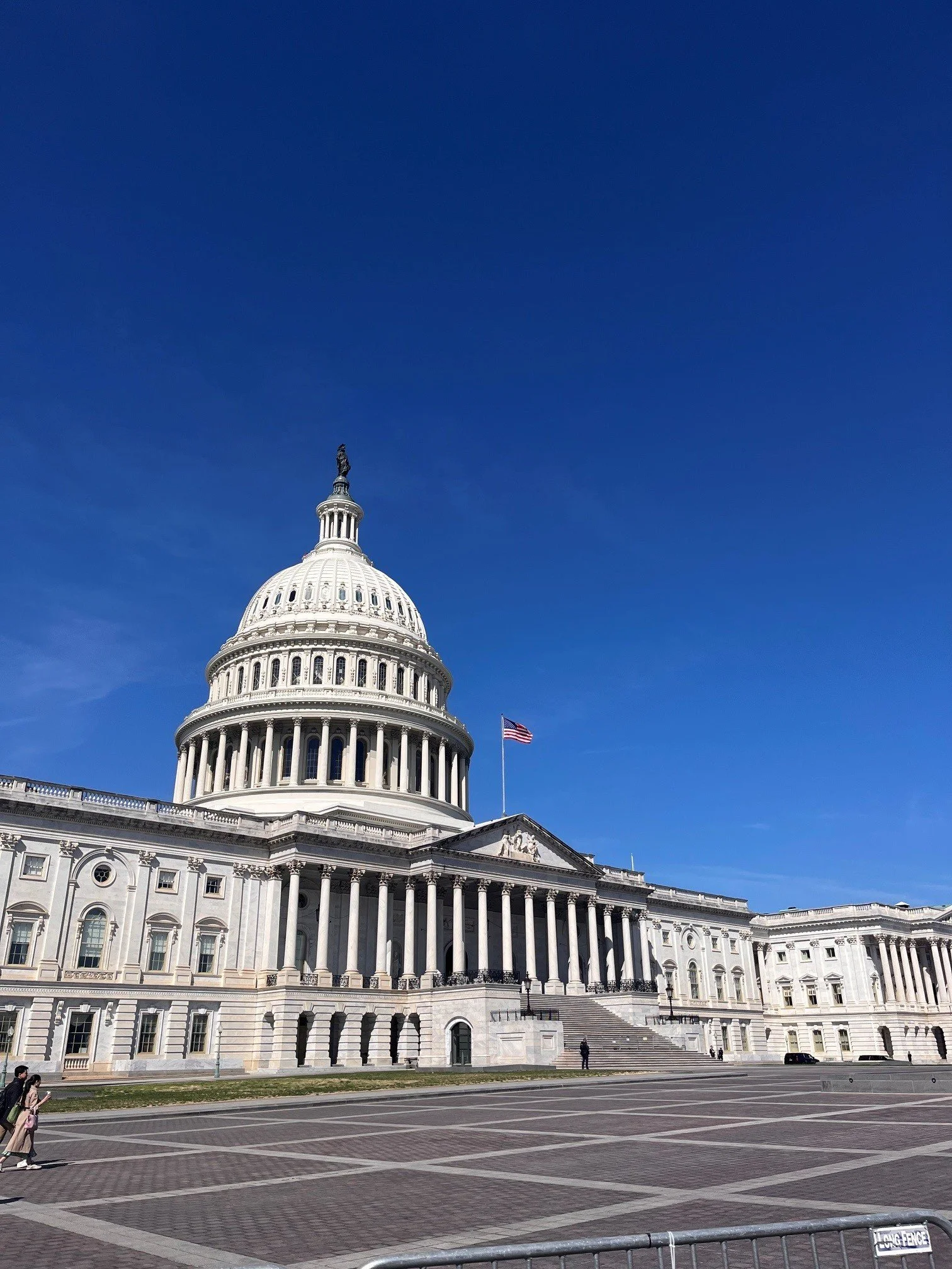 The United States Capitol building under a clear blue sky, with the American flag flying on a pole in front.