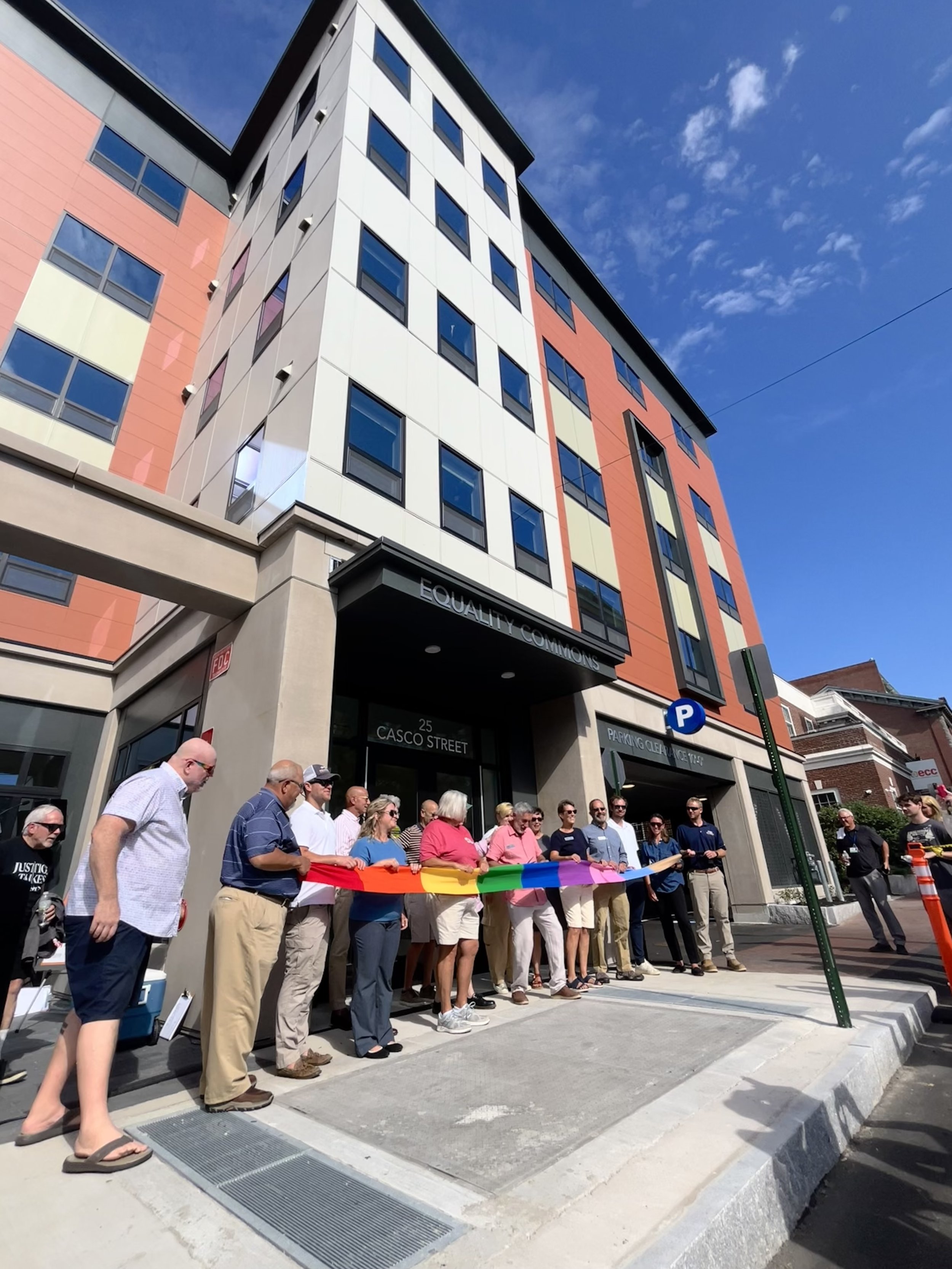 Group of people holding a rainbow pride flag during a public gathering or protest in front of a modern multi-story building.