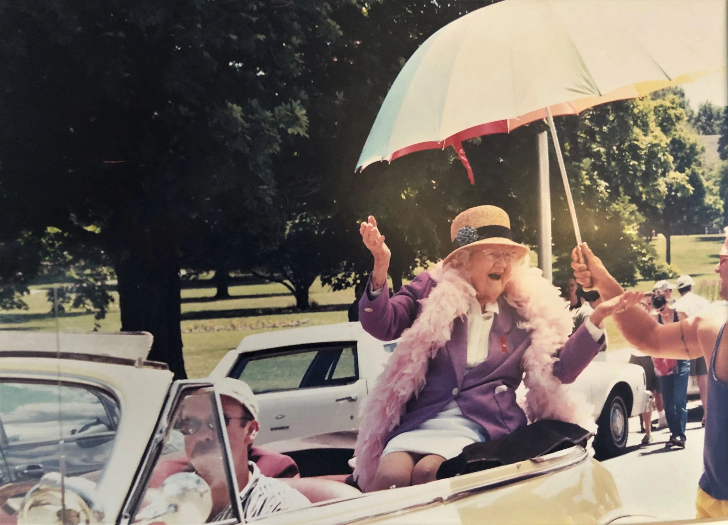 woman wearing a hat and a feather boa riding in a vintage convertible car with other people. She is holding a colorful umbrella at a pride parade  event in a park with trees in the background.