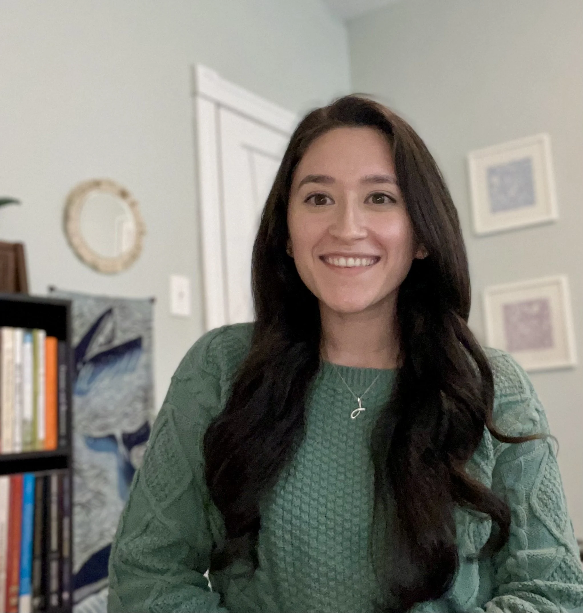 A woman with dark brown hair smiling, wearing a green sweater and a cross necklace, in a room with framed art and a bookshelf with books.