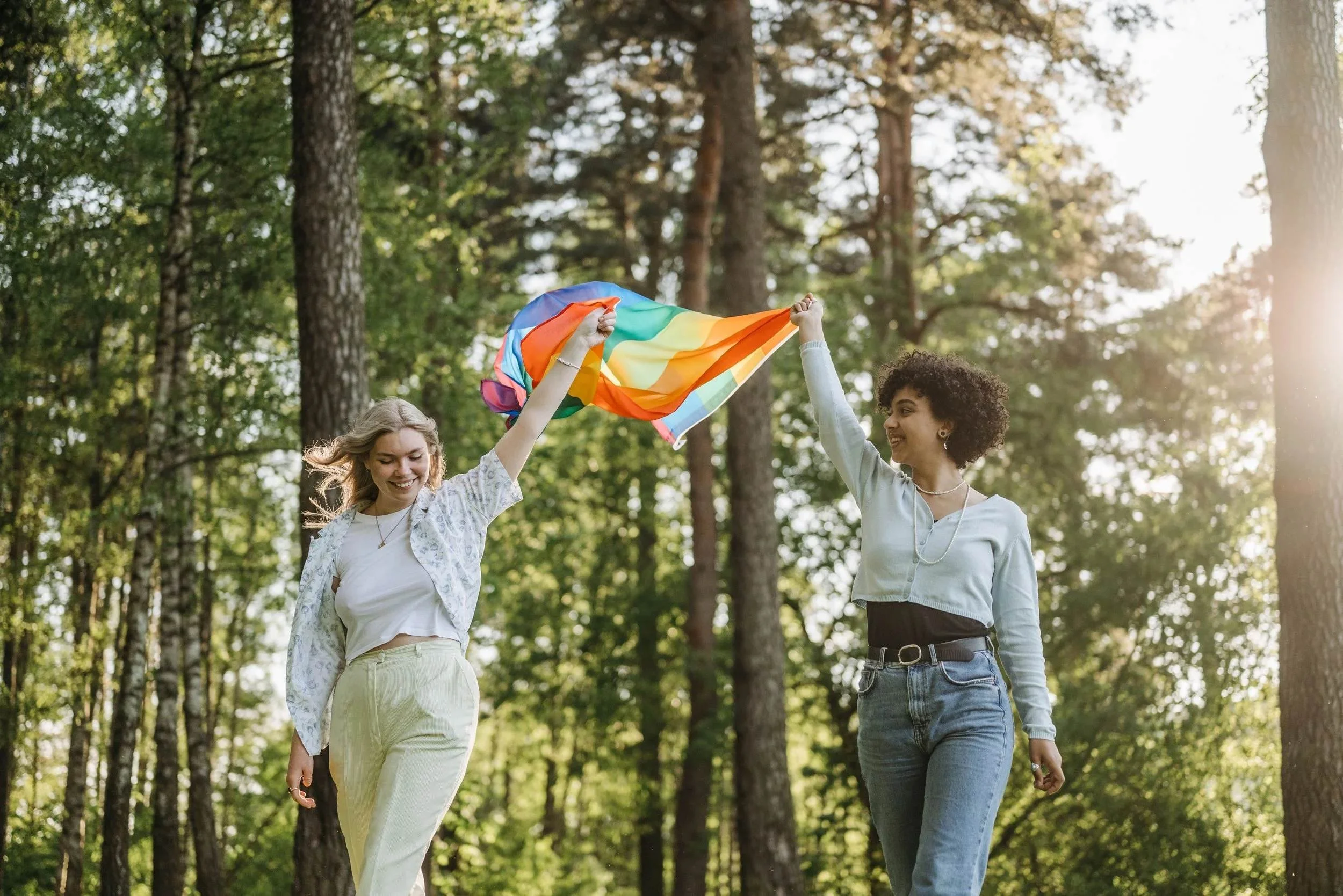 Two women walking together in a forest, one waving a rainbow pride flag, representing couples therapy in Los Angeles