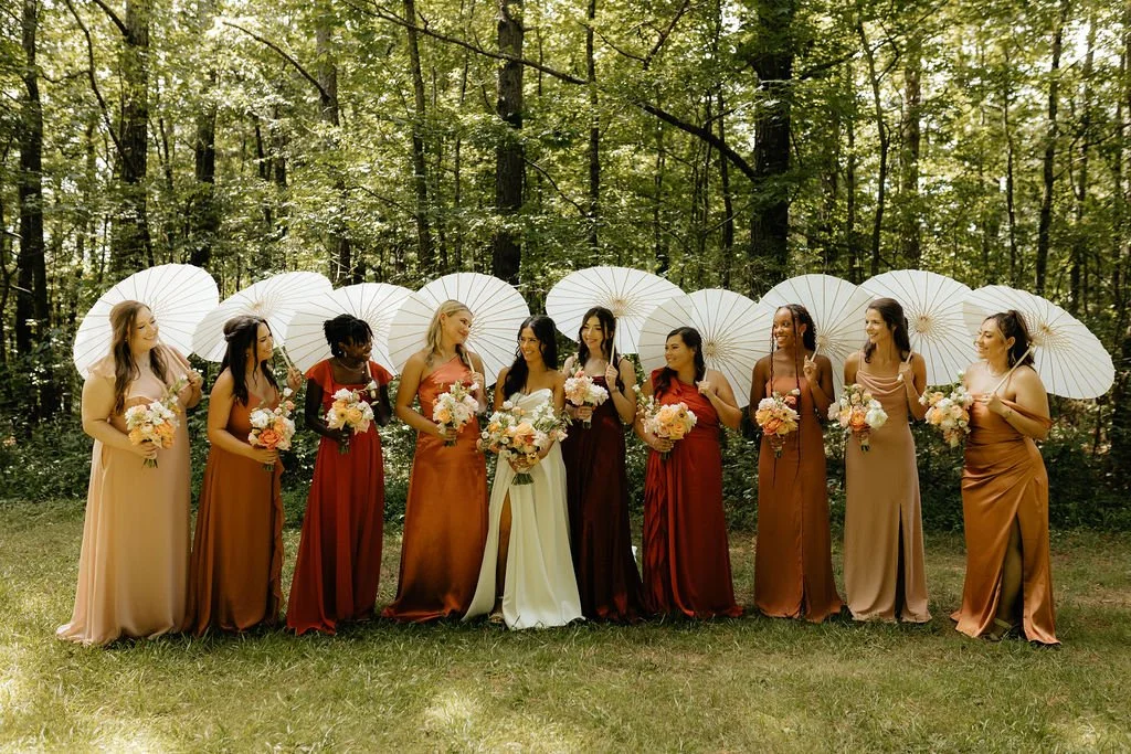 A bride in a white dress standing among nine bridesmaids in dresses of various earth tones, all holding bouquets of flowers, in a forest setting with white parasols in the background.