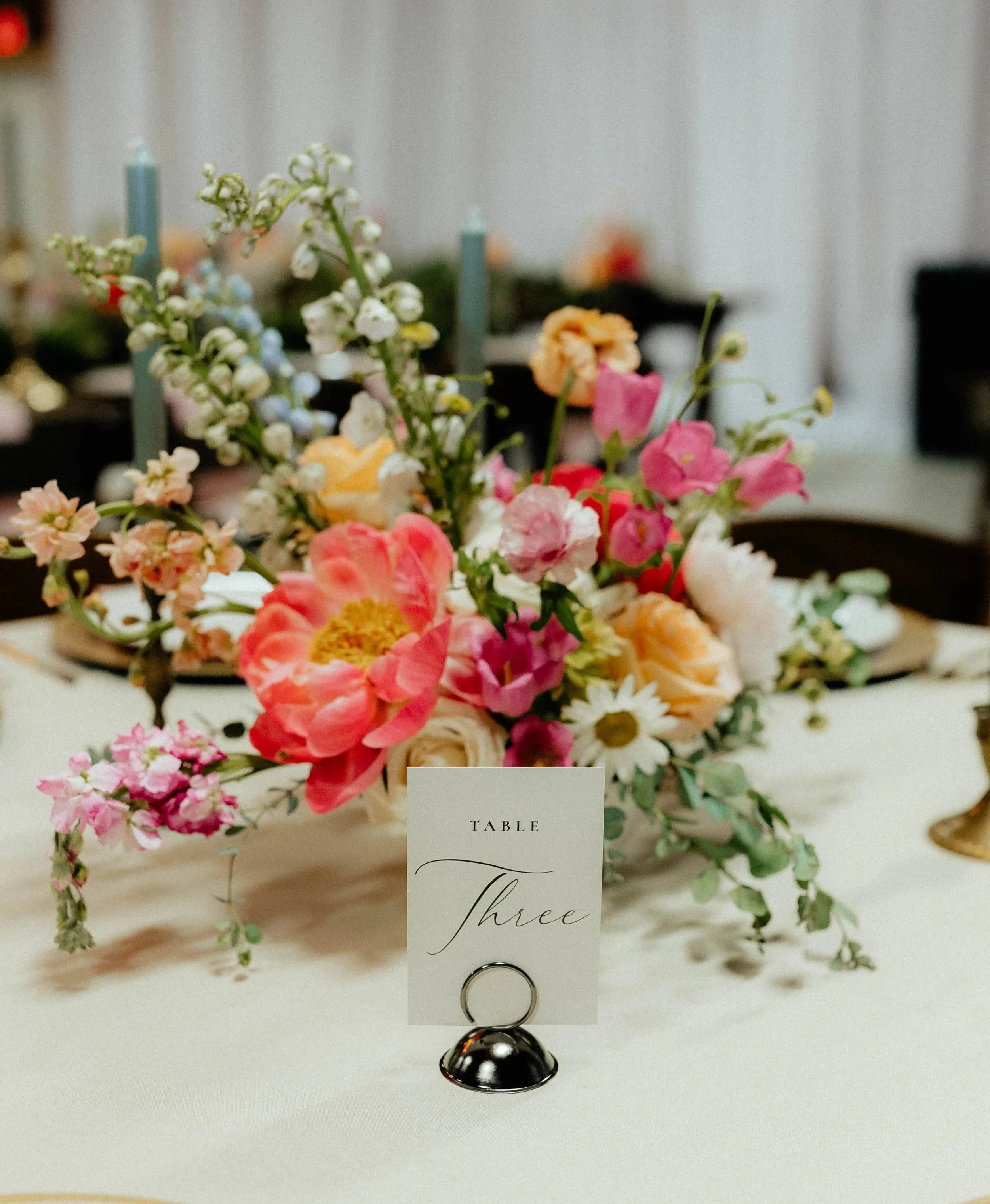 A floral centerpiece on a white table with a small card labeled 'Table Three' and a silver ring holder.