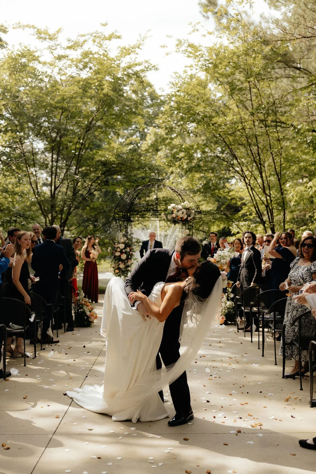 A groom dips and kisses a bride during an outdoor wedding ceremony, surrounded by guests and decorated with flowers and trees.
