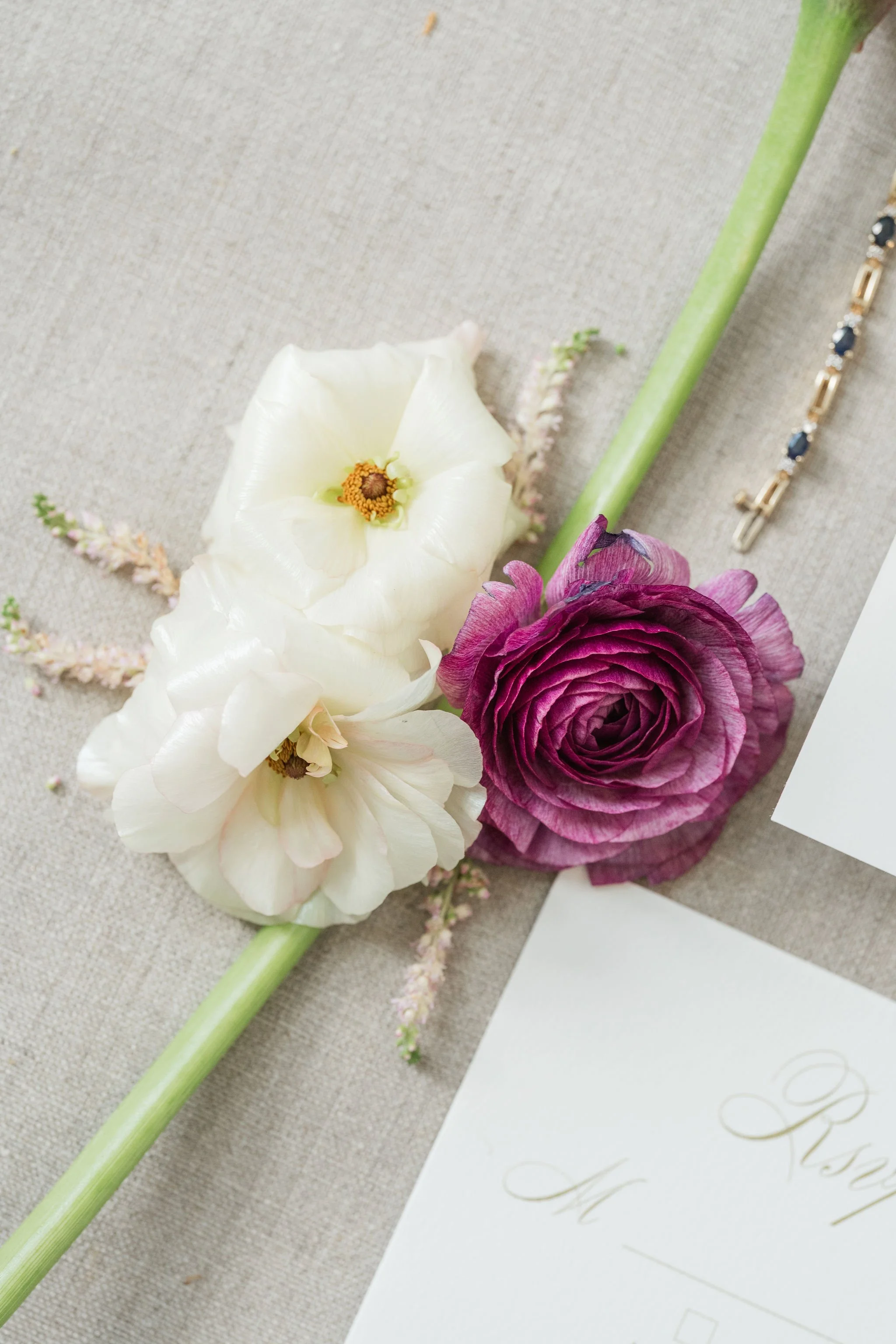Close-up of a floral arrangement with white, purple, and pink flowers, a green stem, and elegant gold and blue jewelry, on a beige fabric surface next to white paper with gold calligraphy.