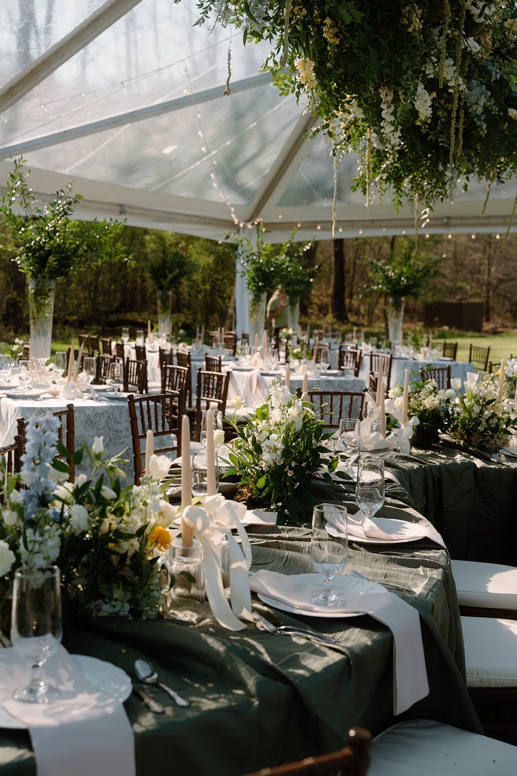 Elegant outdoor wedding reception with round tables and chairs decorated with white floral centerpieces under a white tent with hanging greenery and string lights.
