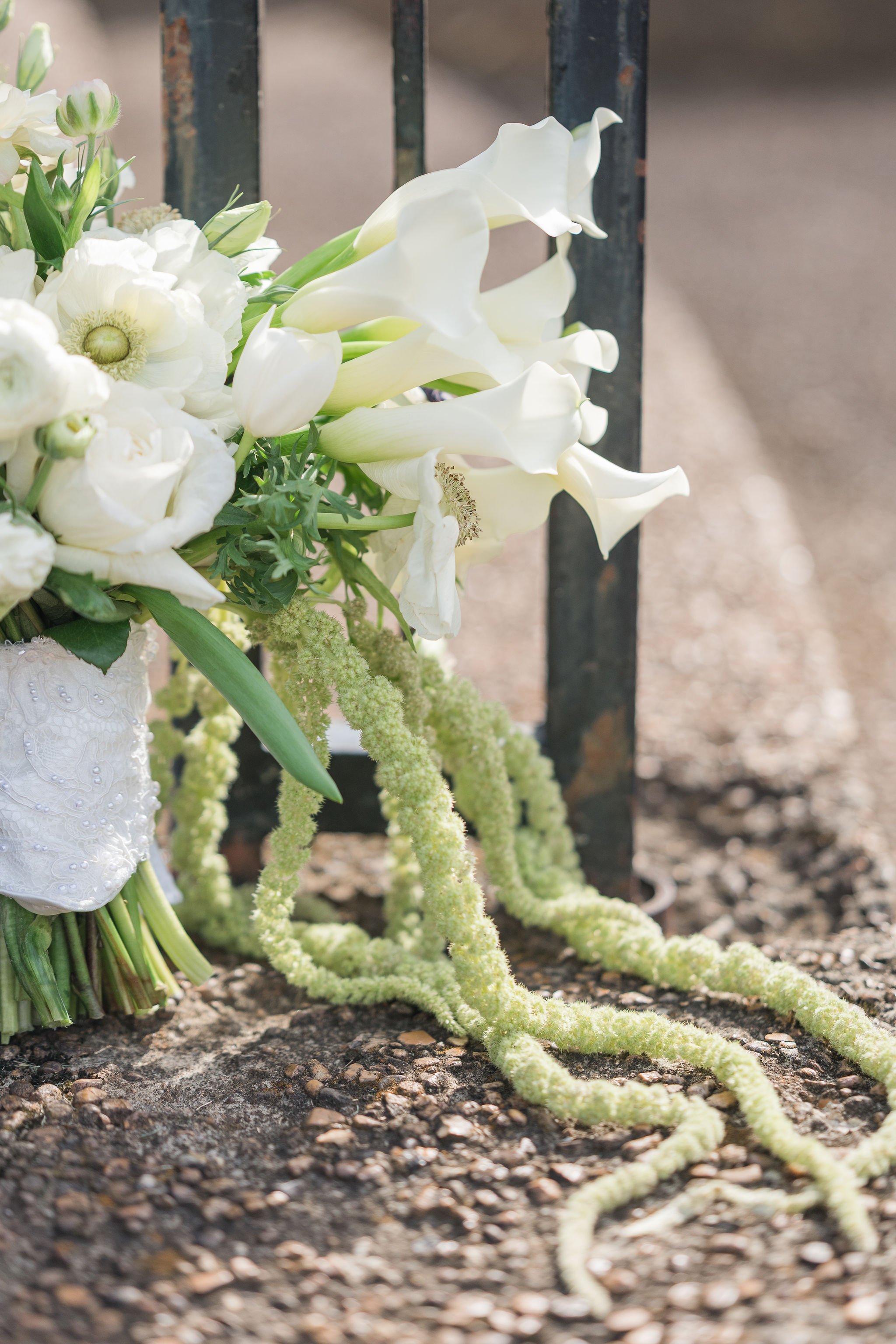 White floral arrangement with calla lilies, lisianthus, and greenery placed on the ground near a black iron fence post, outdoors.