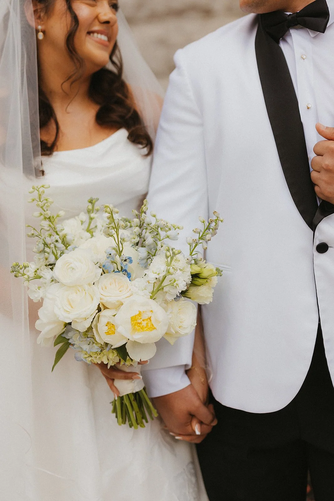 A bride and groom holding hands at their wedding, with the bride holding a bouquet of white and pale yellow flowers, including roses and peonies, while smiling at each other.
