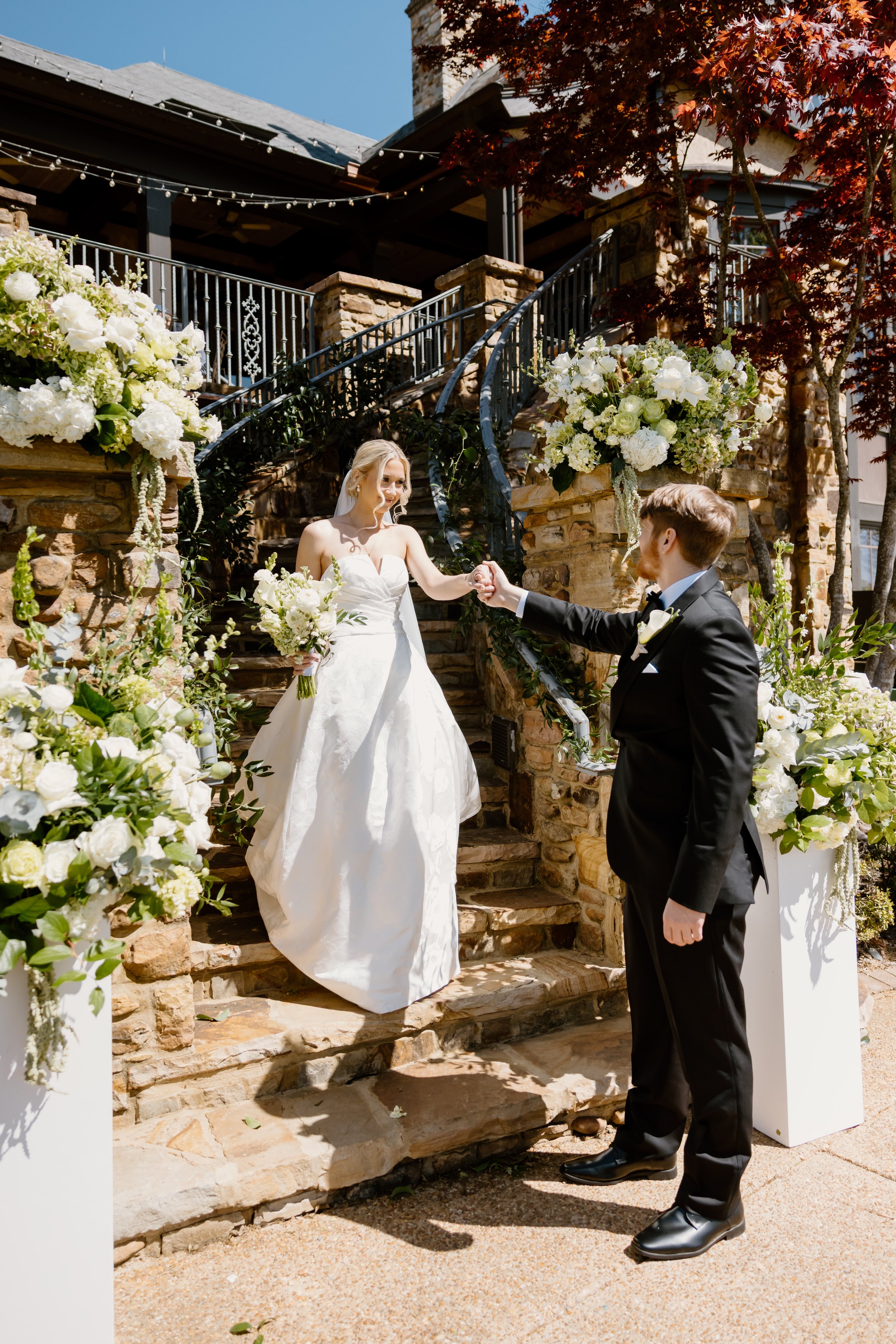 A bride in a white wedding dress holding a bouquet and smiling at a groom in a black tuxedo, who is holding her hand during a wedding ceremony outdoors on stone stairs decorated with white flowers and greenery.
