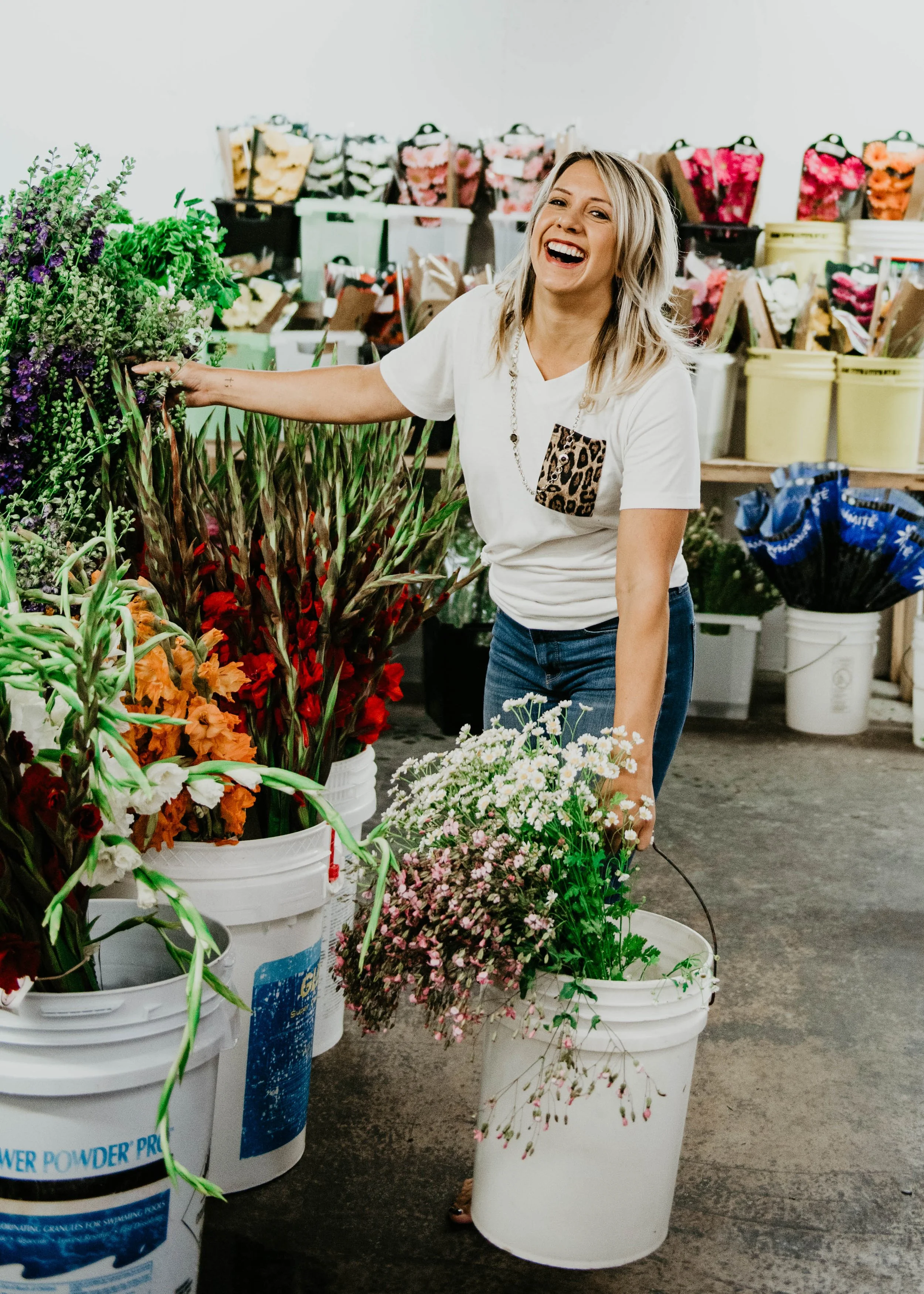 A woman in a white t-shirt with a leopard print pocket is smiling and holding a white bucket of pink flowers in a store with buckets of flowers and gardening supplies.