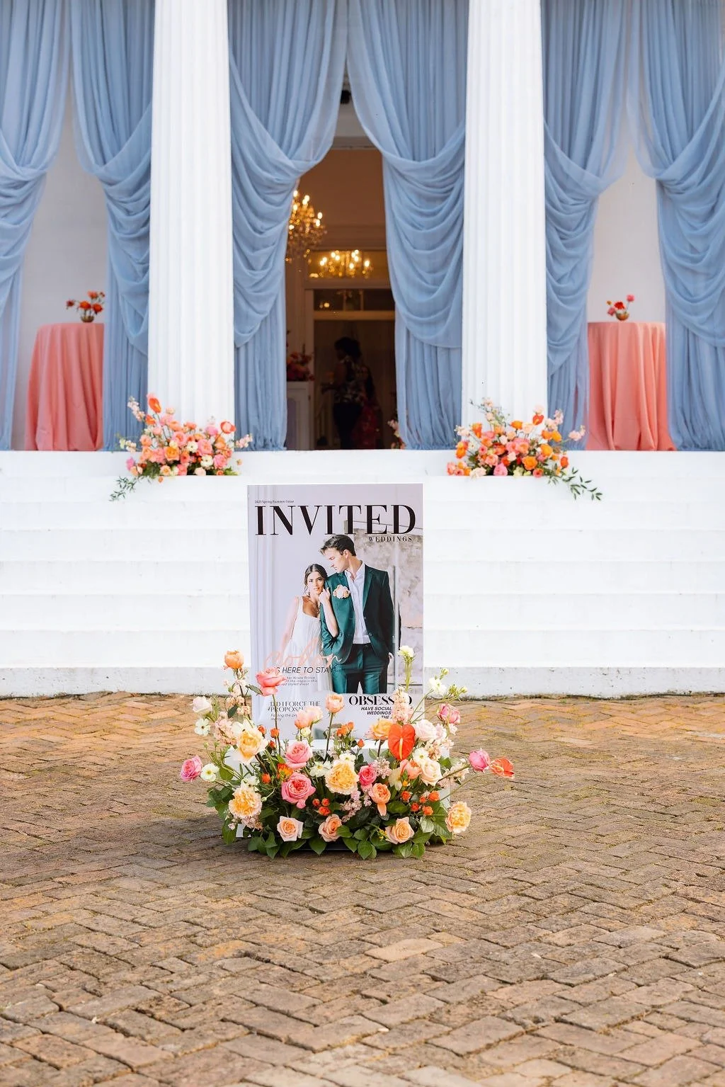 Wedding invitation sign featuring a picture of a bride and groom, surrounded by pink and peach flowers, placed in front of a white staircase with blue curtains and floral arrangements.