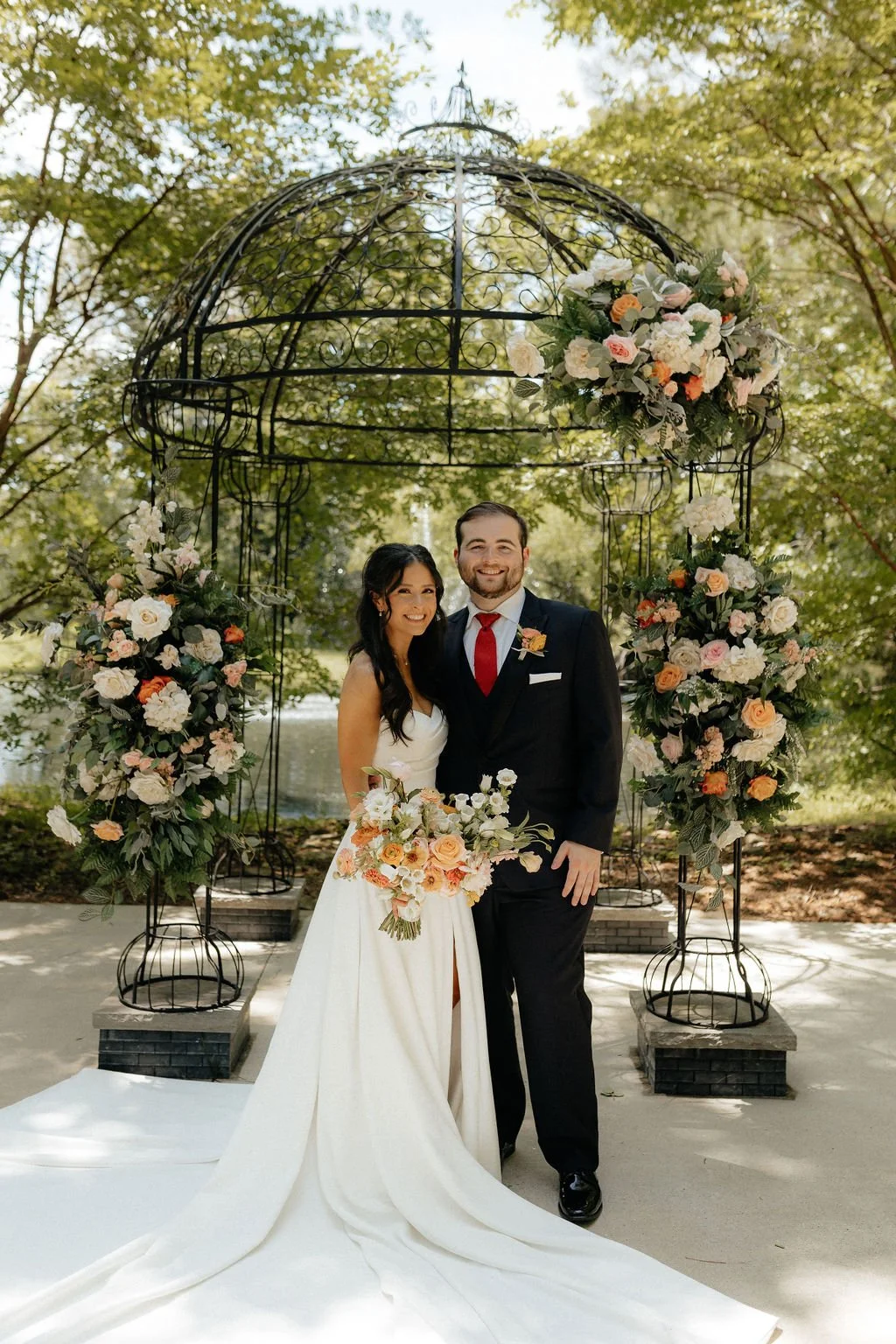 A bride and groom smiling and posing for a wedding photo outdoors under a metal arbor decorated with pink, white, and peach flowers.