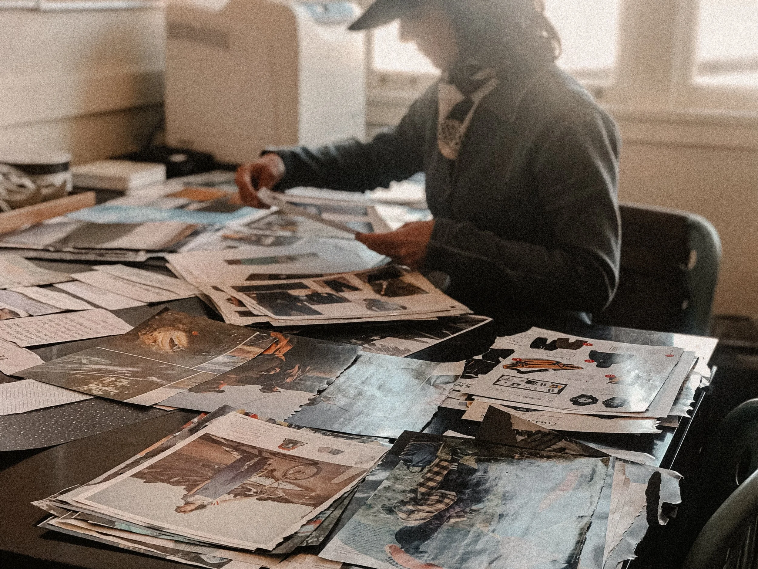 Designer sorting through stacks of artwork, photographs, and papers spread across a table for trend research.