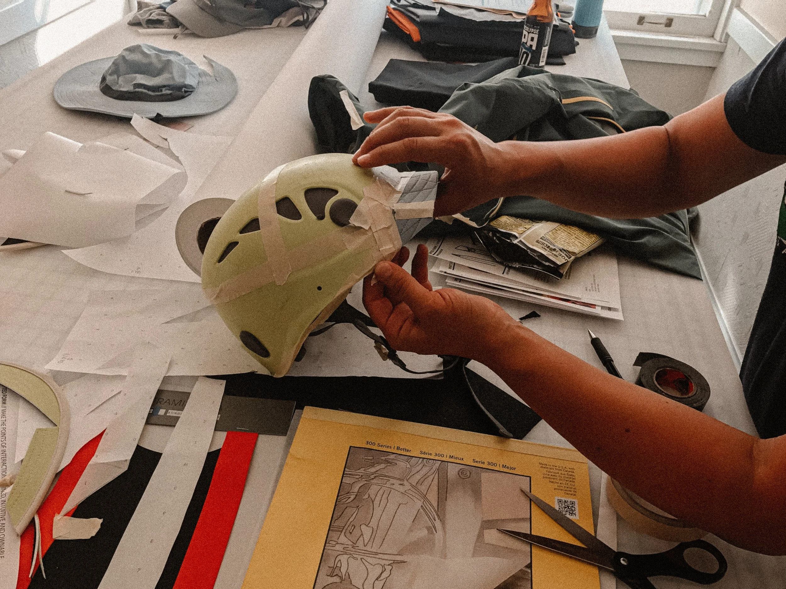 Person taping a light green helmet on a craft-covered worktable surrounded by scissors, paper, fabric, and art supplies
