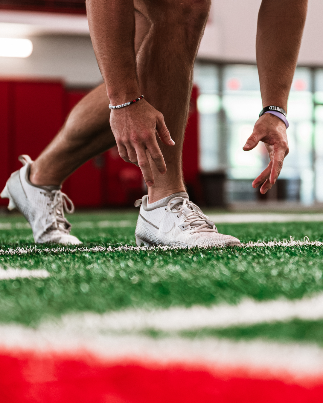 An athlete in white cleats crouches at the starting line on a green indoor track, preparing for a race.
