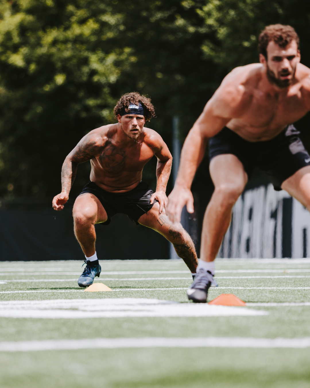 Two shirtless athletes with athletic builds doing agility drills on a football field with orange cones, surrounded by trees.