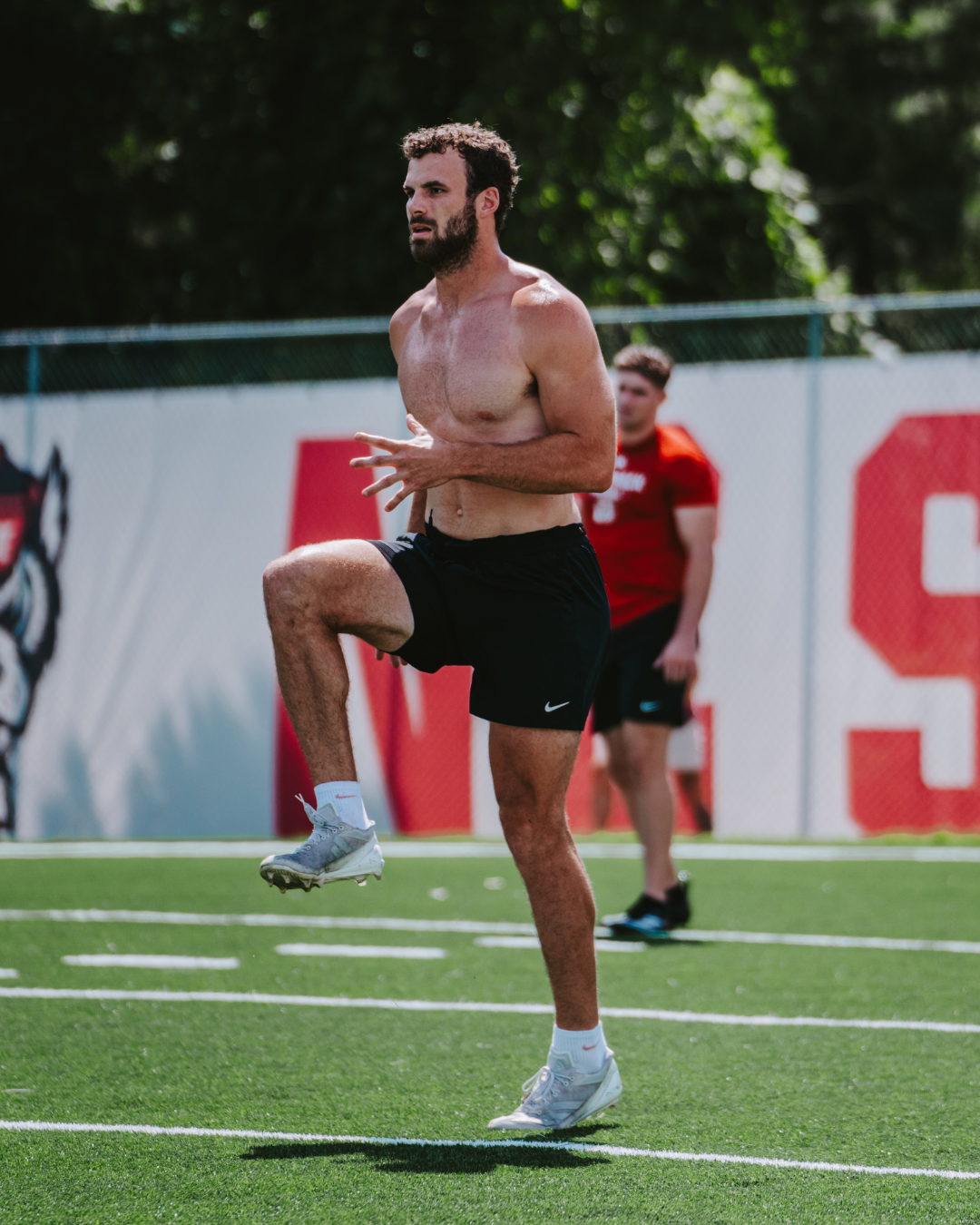 A shirtless athlete in black shorts and white sneakers exercises on a green field, lifting his right knee with his hands near his stomach. Performing a sprinting drill. Performance Training for athletes