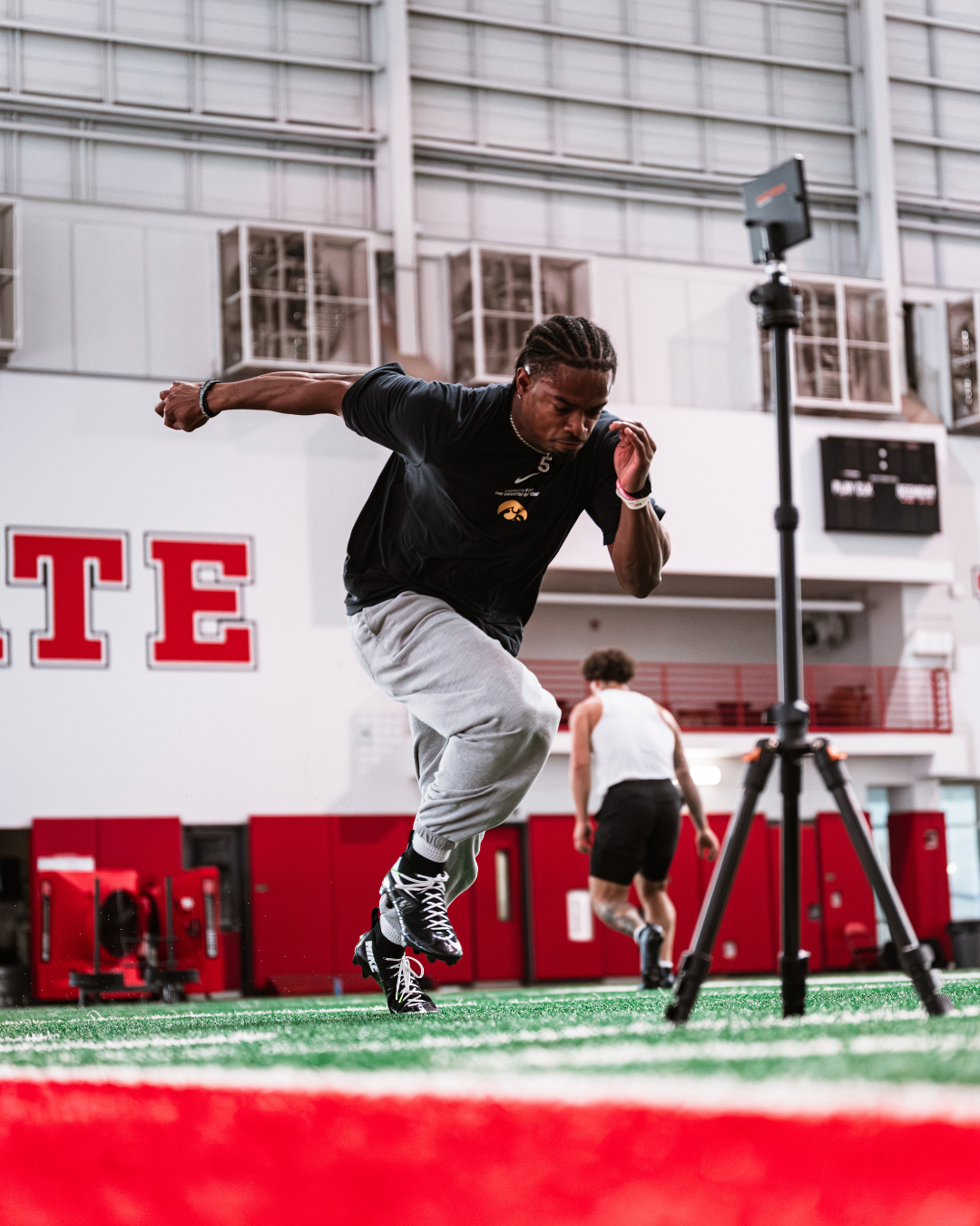 Athlete sprinting inside a sports facility, sport performance training with a person in the background and in NC State University facility with red and white walls.