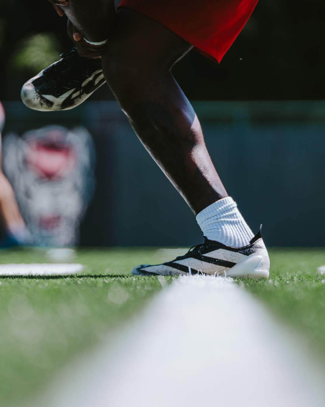 Close-up of an athlete in black and white cleats and white socks, bent over on a green field, with a blurred mascot or logo in the background.