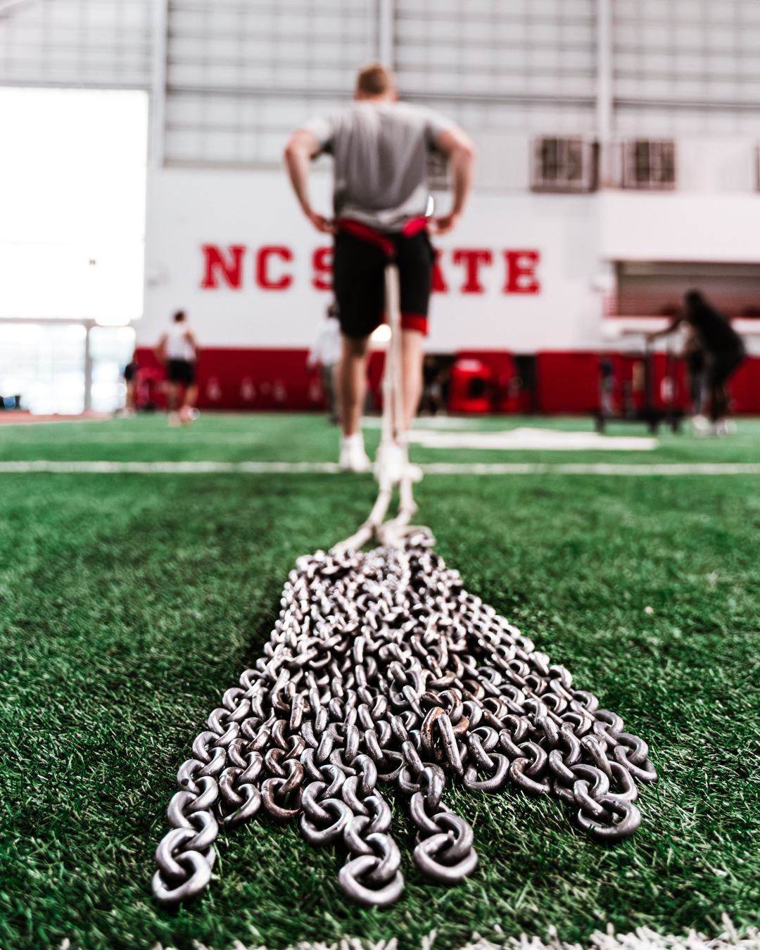 A person standing on an indoor college facility, pulling a long chain along the turf at North Carolina State University, with other individuals practicing in the background.