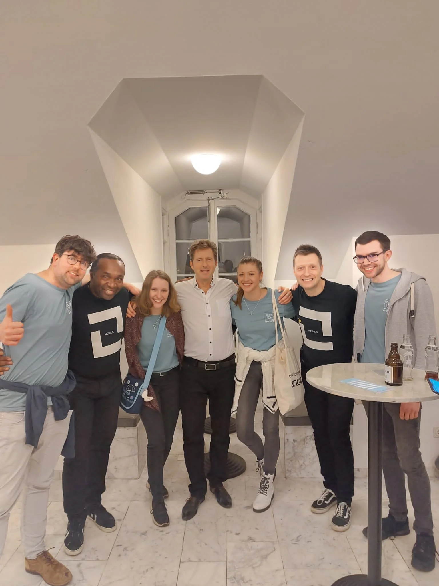 Group of eight diverse young adults smiling and posing together indoors, some with arms around each other's shoulders.