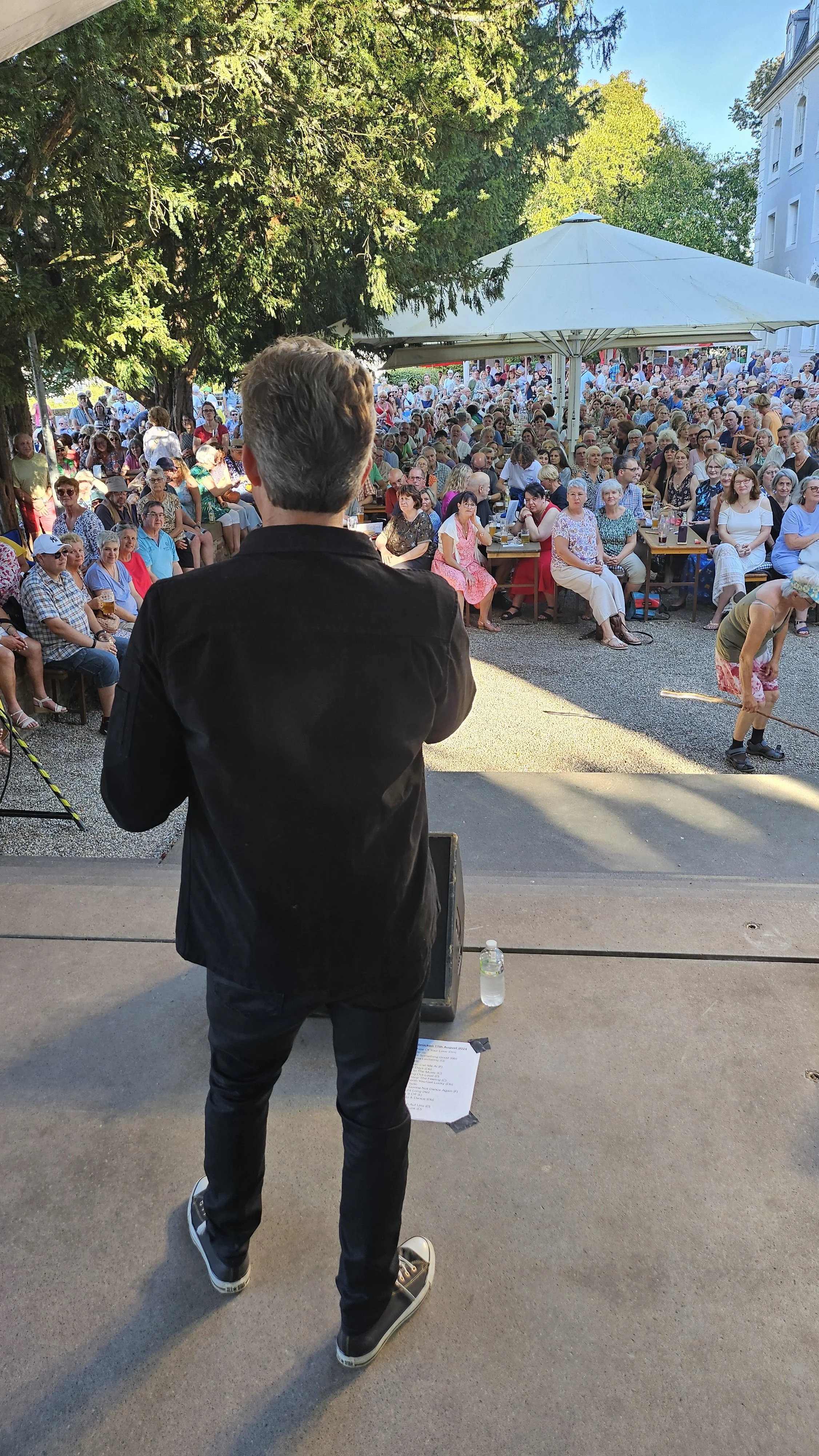 A man with gray hair dressed in black stands on stage facing a large outdoor audience seated at picnic tables under large white umbrellas. The audience is a diverse group of adults enjoying an event in a park-like setting with green trees and a blue 