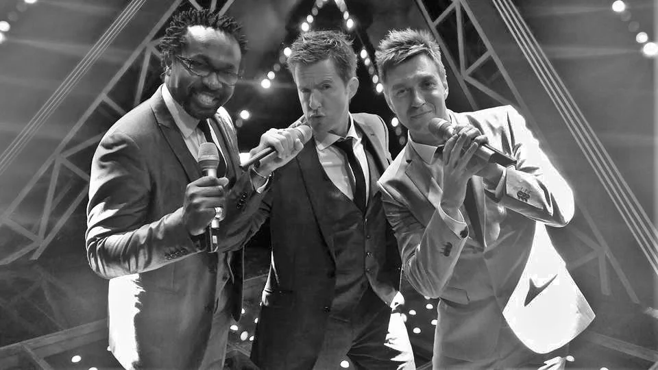 Three men in suits holding microphones on a stage with a lit arch and spotlights behind them.