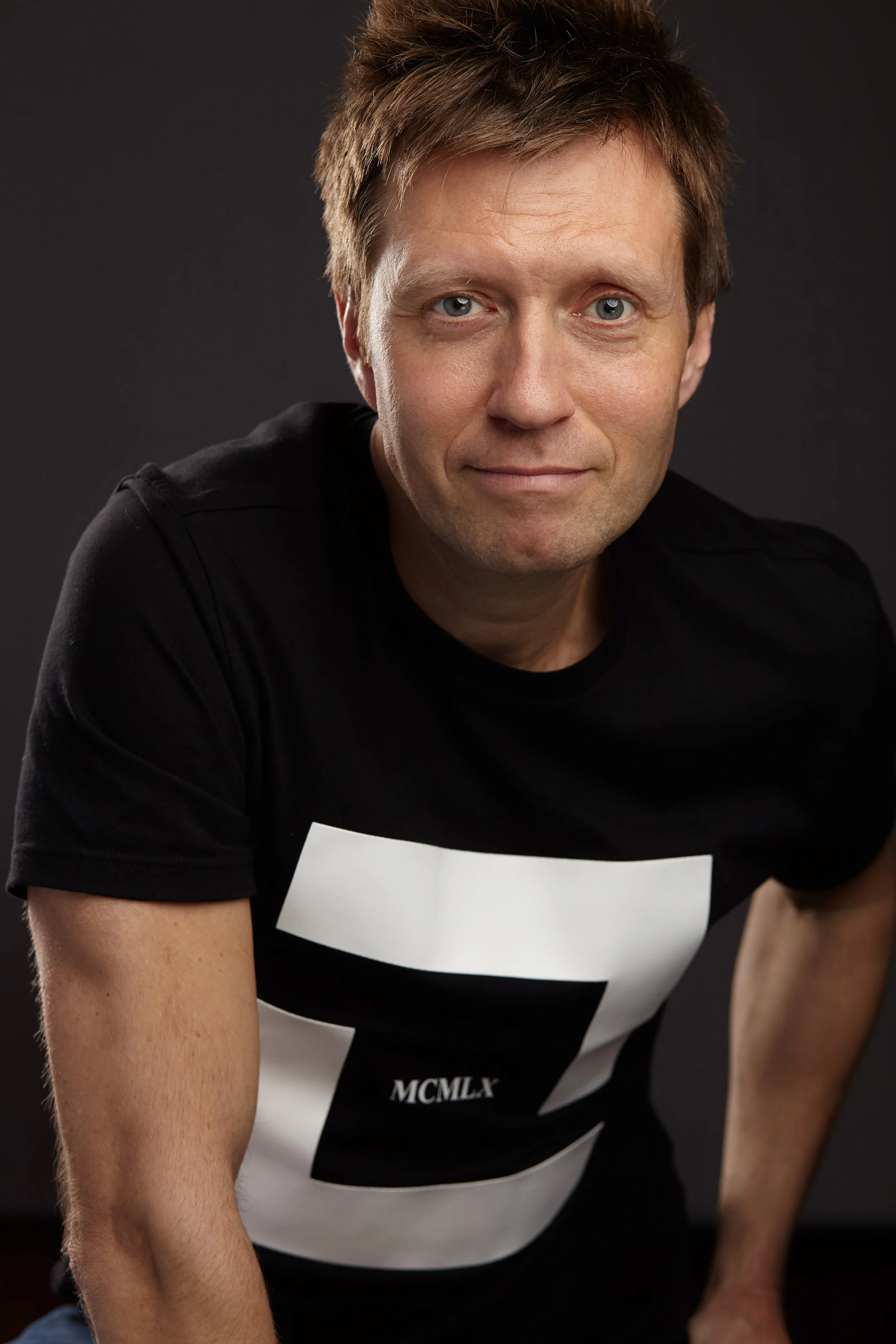 Close-up of a man with short brown hair and blue eyes, wearing a black and white striped t-shirt with the Roman numerals MCMLX, against a black background.