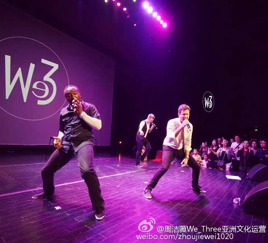 Three male singers performing on a stage with purple lighting, singing into microphones, with a group of people watching in the background, and a large screen with a W3 logo projected on it.