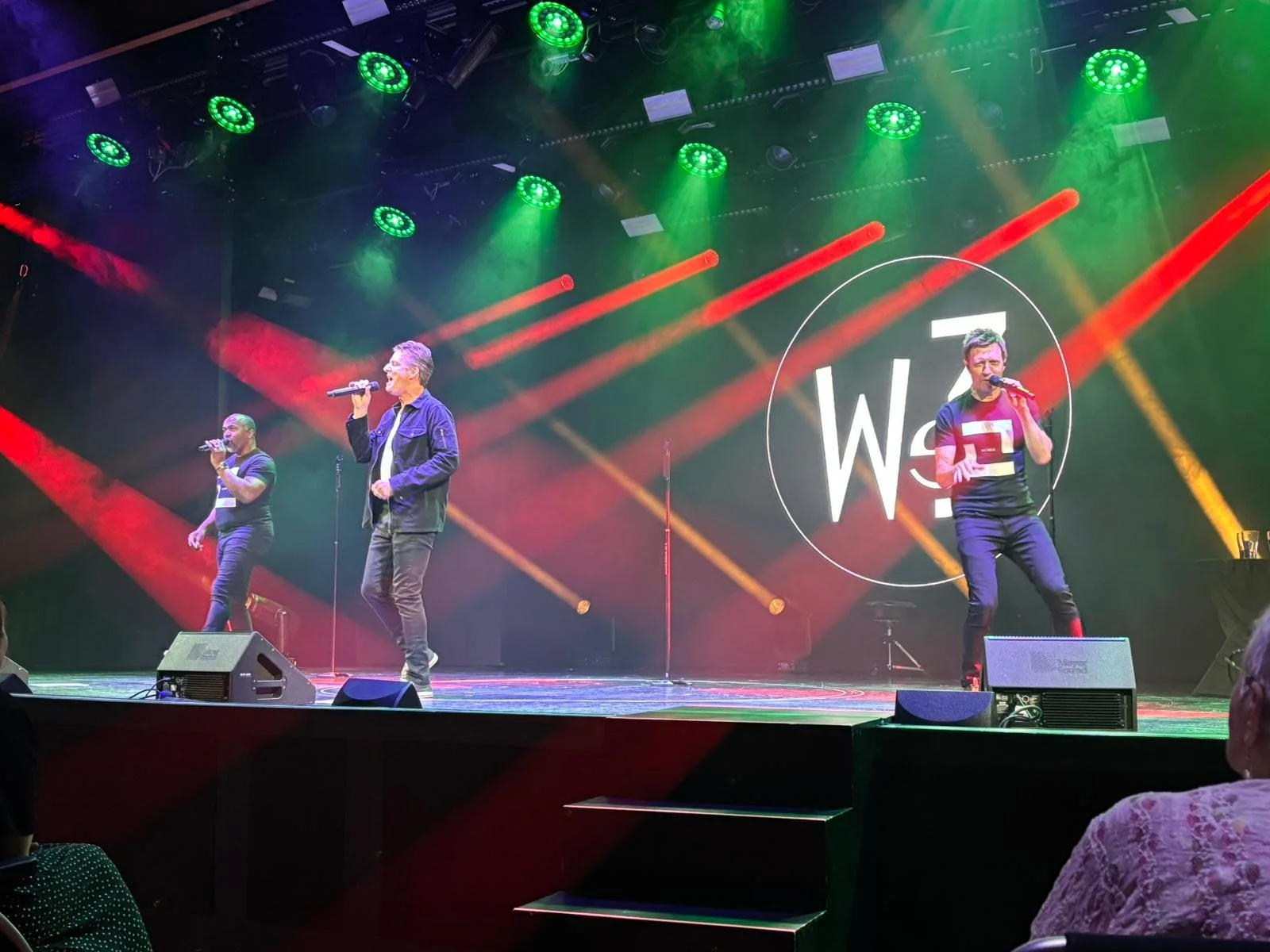 Three performers singing on a stage with colorful lighting and a large backdrop displaying the WSI logo.