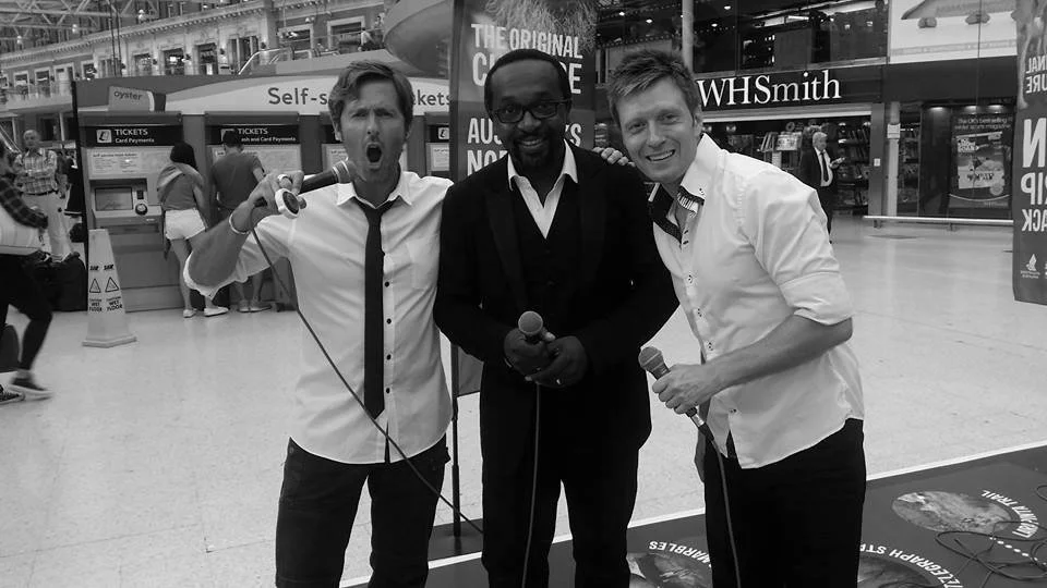 Three men standing in an airport terminal, holding microphones. The man on the left appears to be singing passionately, the man in the middle is smiling, and the man on the right is also smiling. There are people and signs in the background, includin
