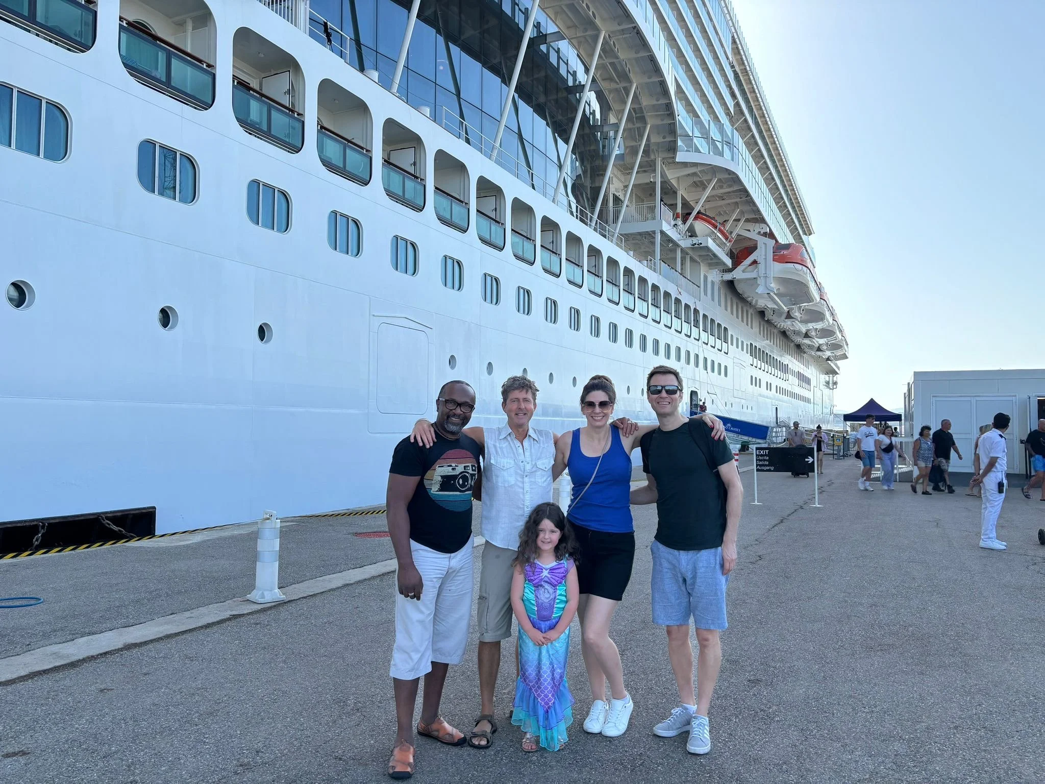 A group of five people, three adults and two children, standing together on a dock in front of a large white cruise ship with multiple decks and rows of windows. The group is smiling, and the children are dressed in colorful costumes, with one girl i