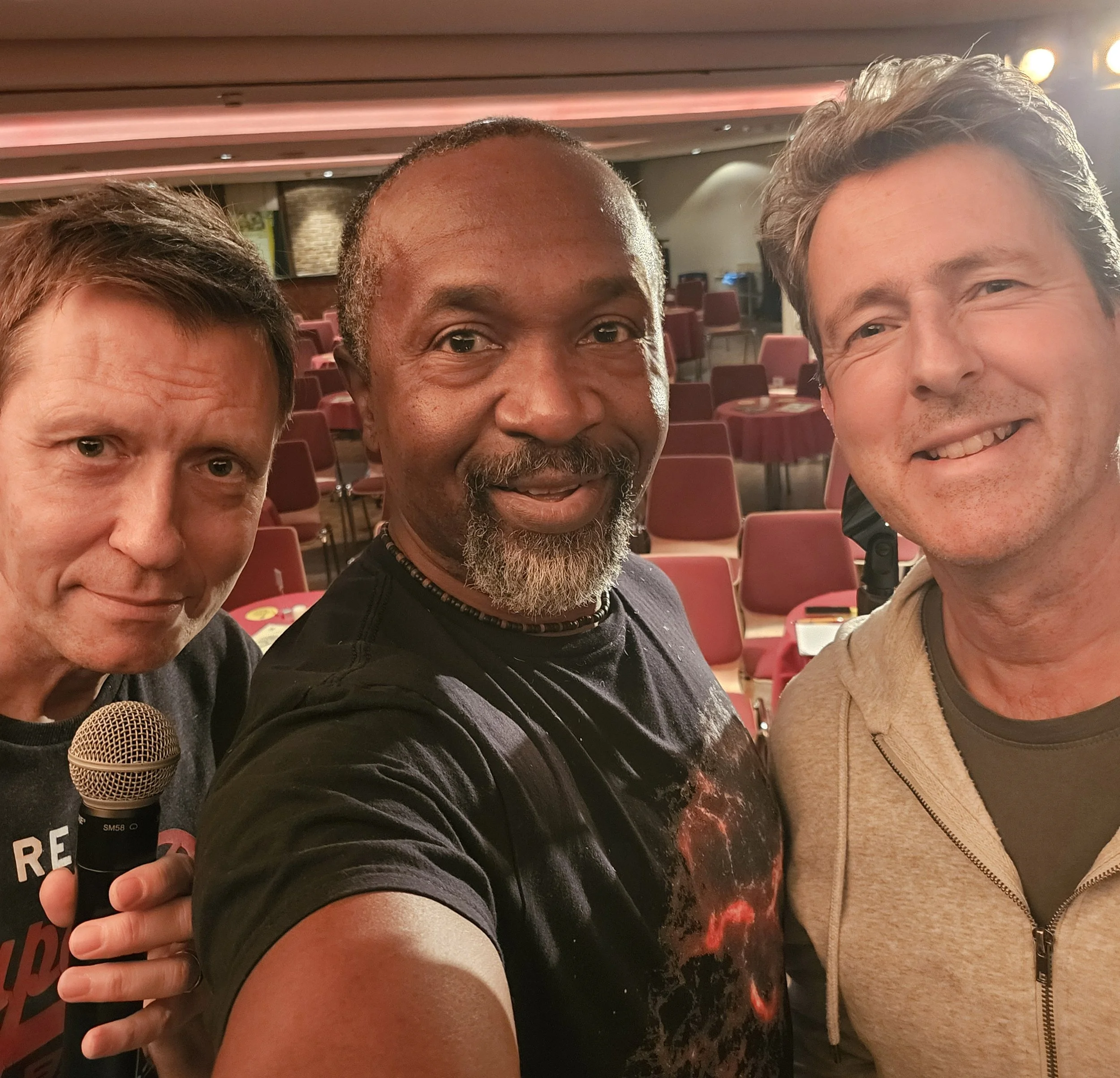 Three men taking a selfie in a restaurant or banquet hall with red chairs and tables in the background.