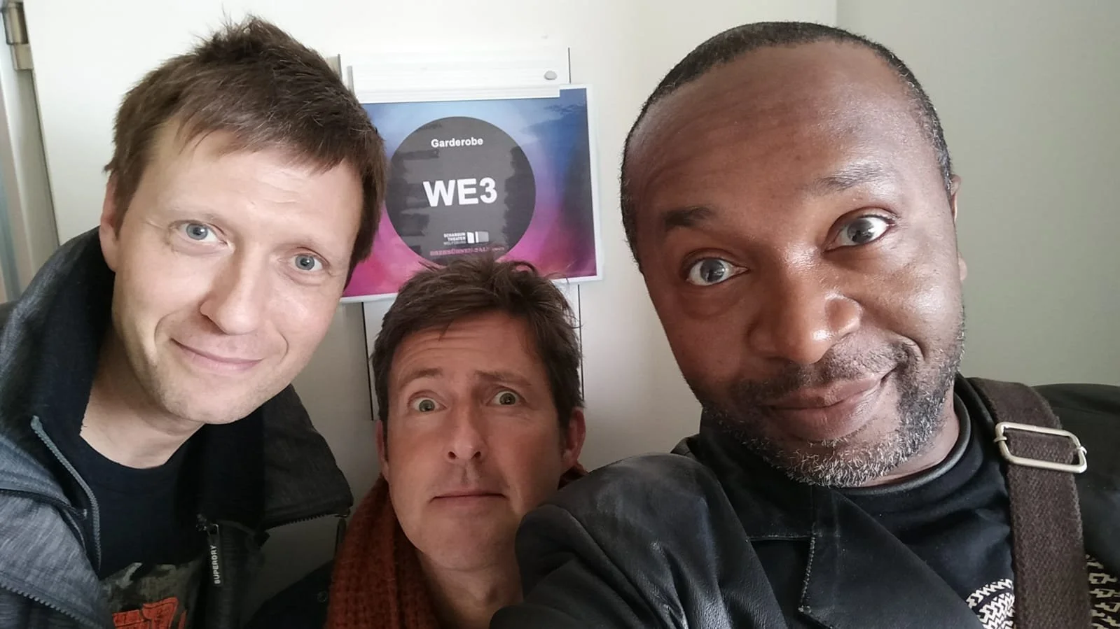 Three men taking a selfie in an airport or train station. Behind them is a sign with a purple and pink design that reads "Garderobe WE3."