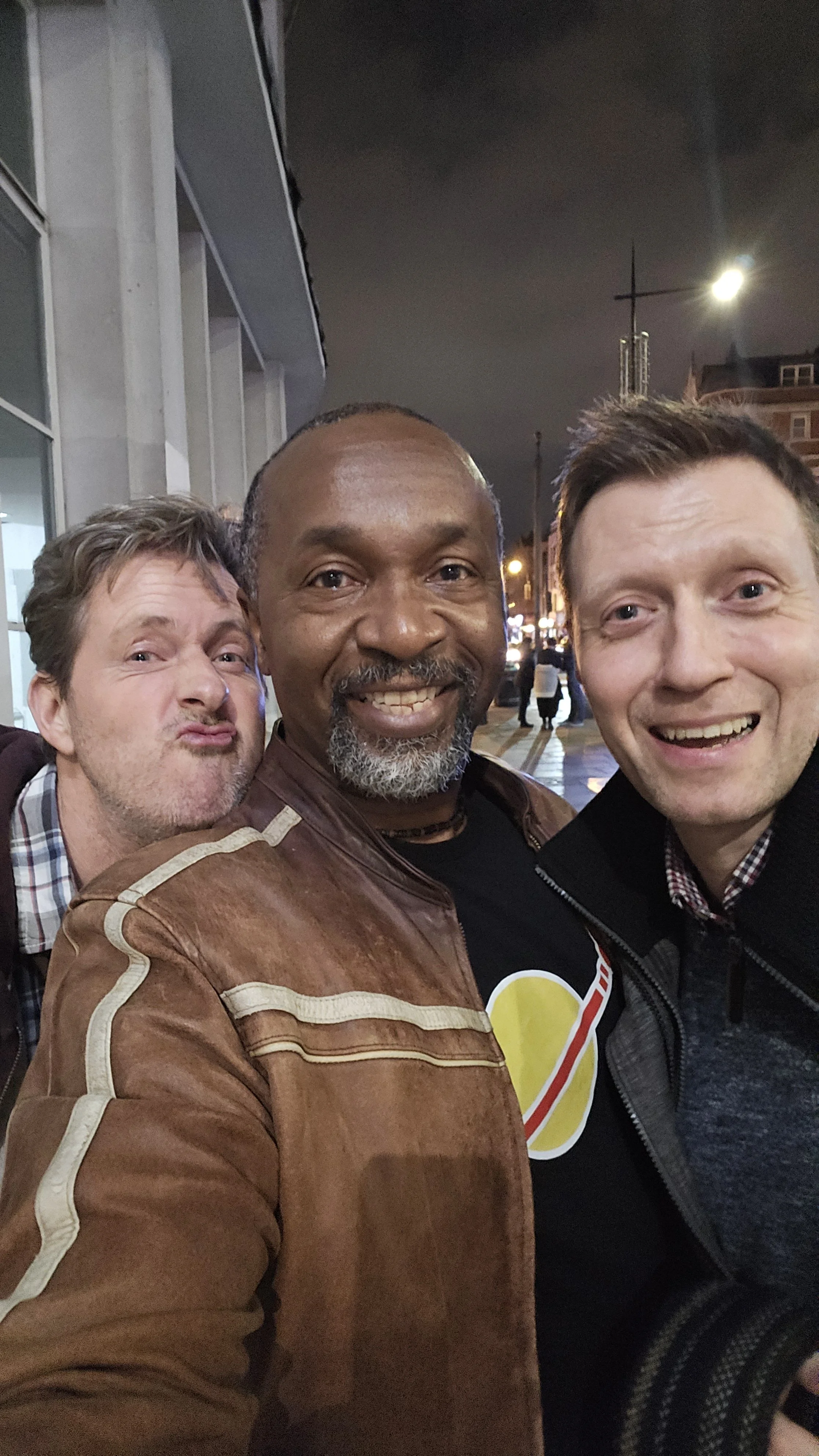 Three men taking a selfie on a city street at night, smiling and posing closely together.